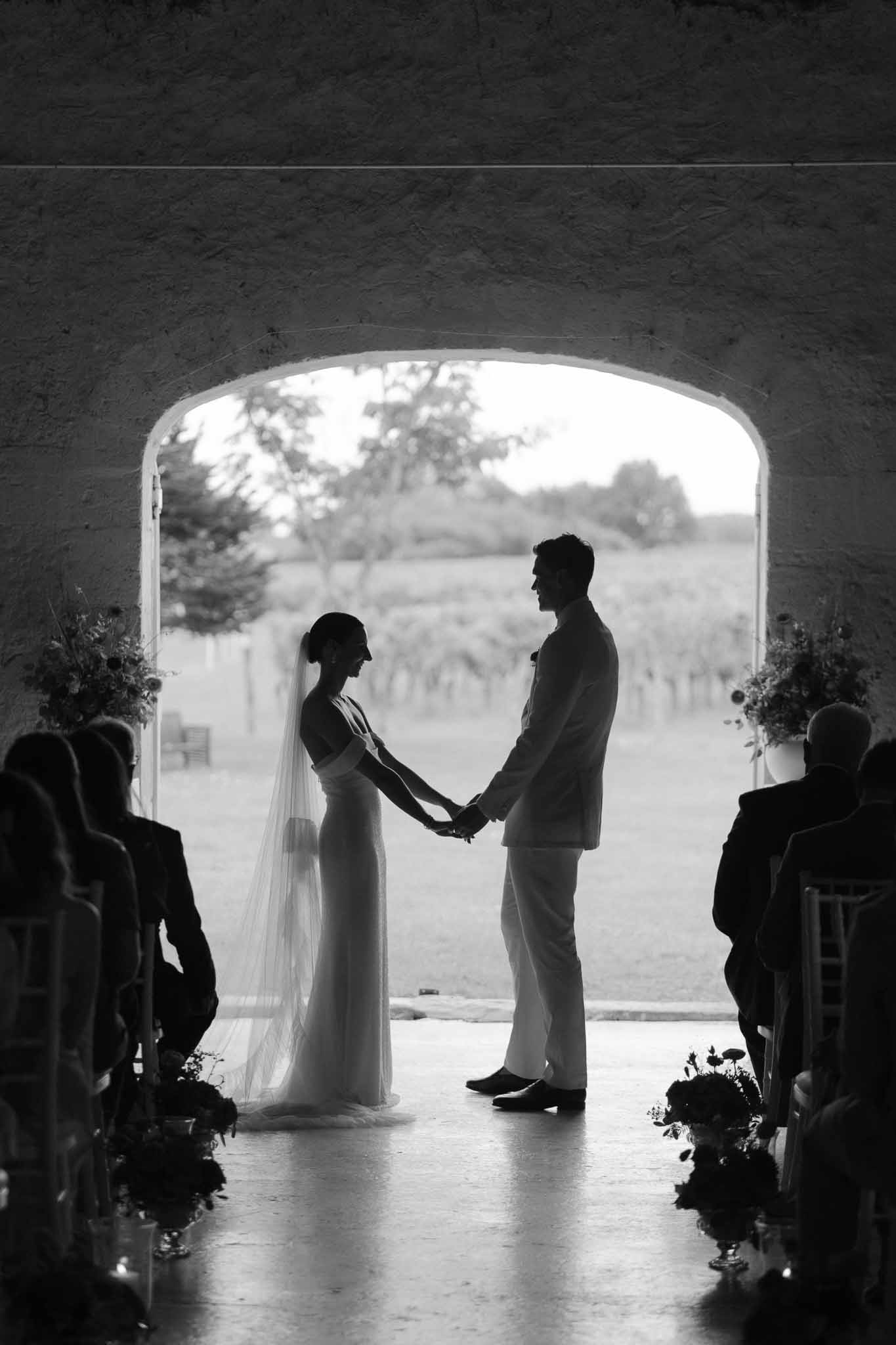 Black-and-white silhouette of couple exchanging vows under a large arch with vineyard backdrop and seated guests