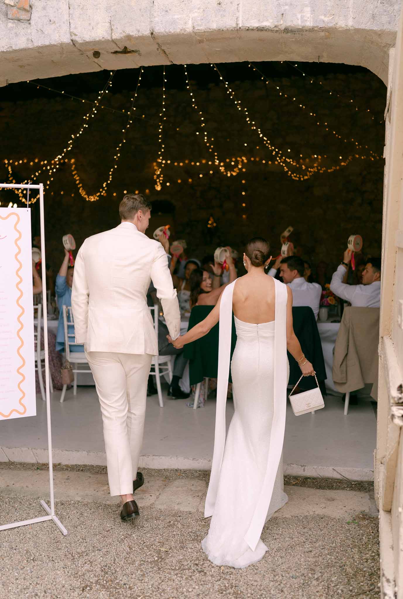 Bride and groom making their entrance into a stone-walled reception venue draped with fairy lights as guests cheer