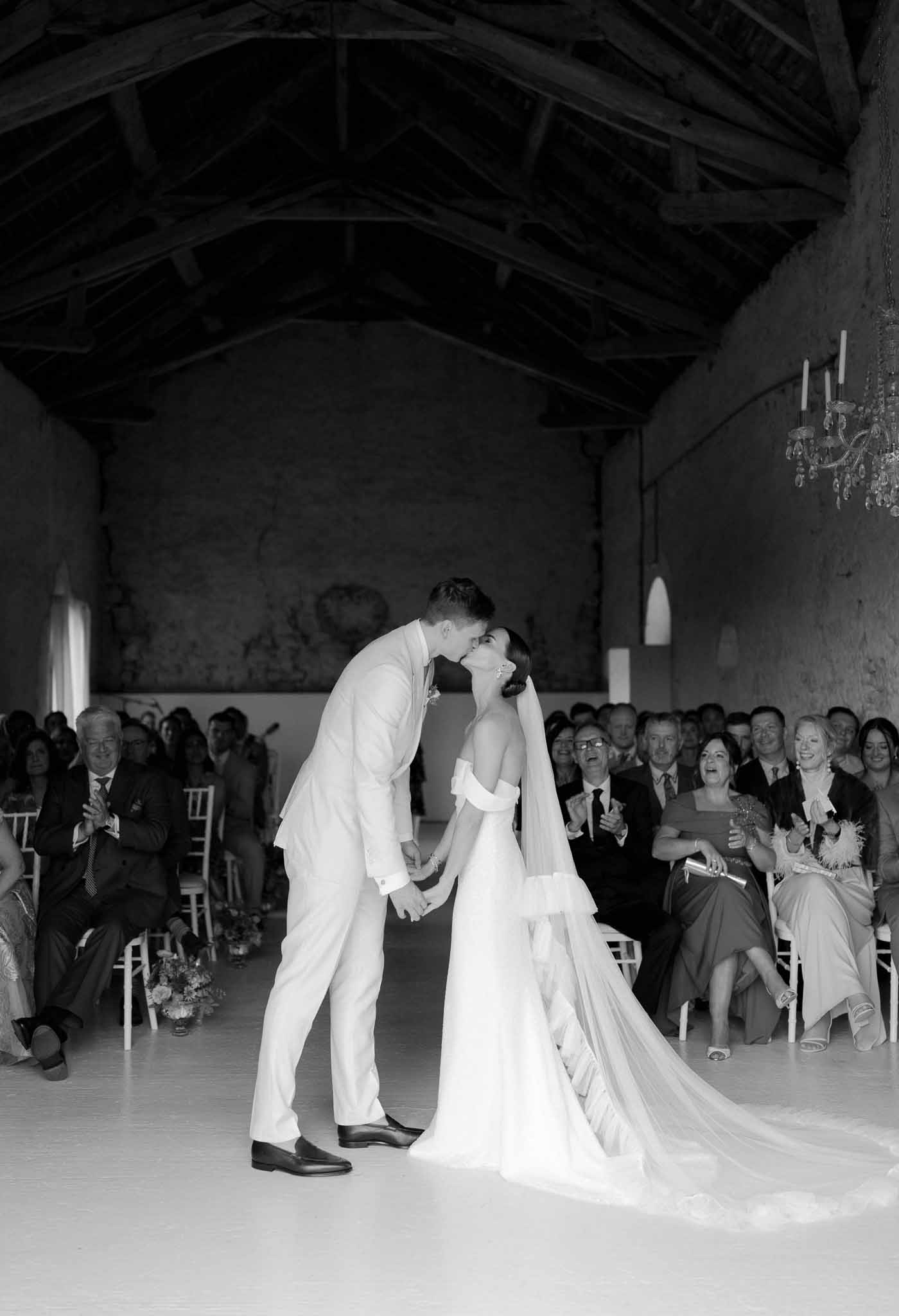 Black-and-white wide shot of couple sharing first kiss in rustic barn ceremony with guests applauding