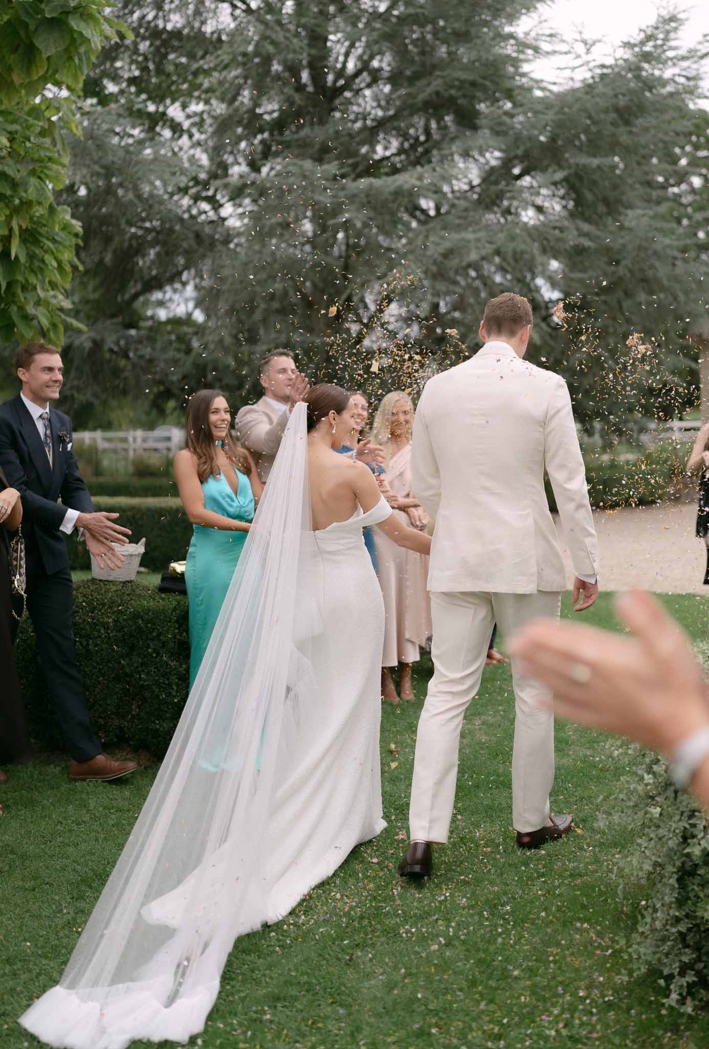 Rear view of couple during gold confetti exit with cathedral veil trailing across estate lawn