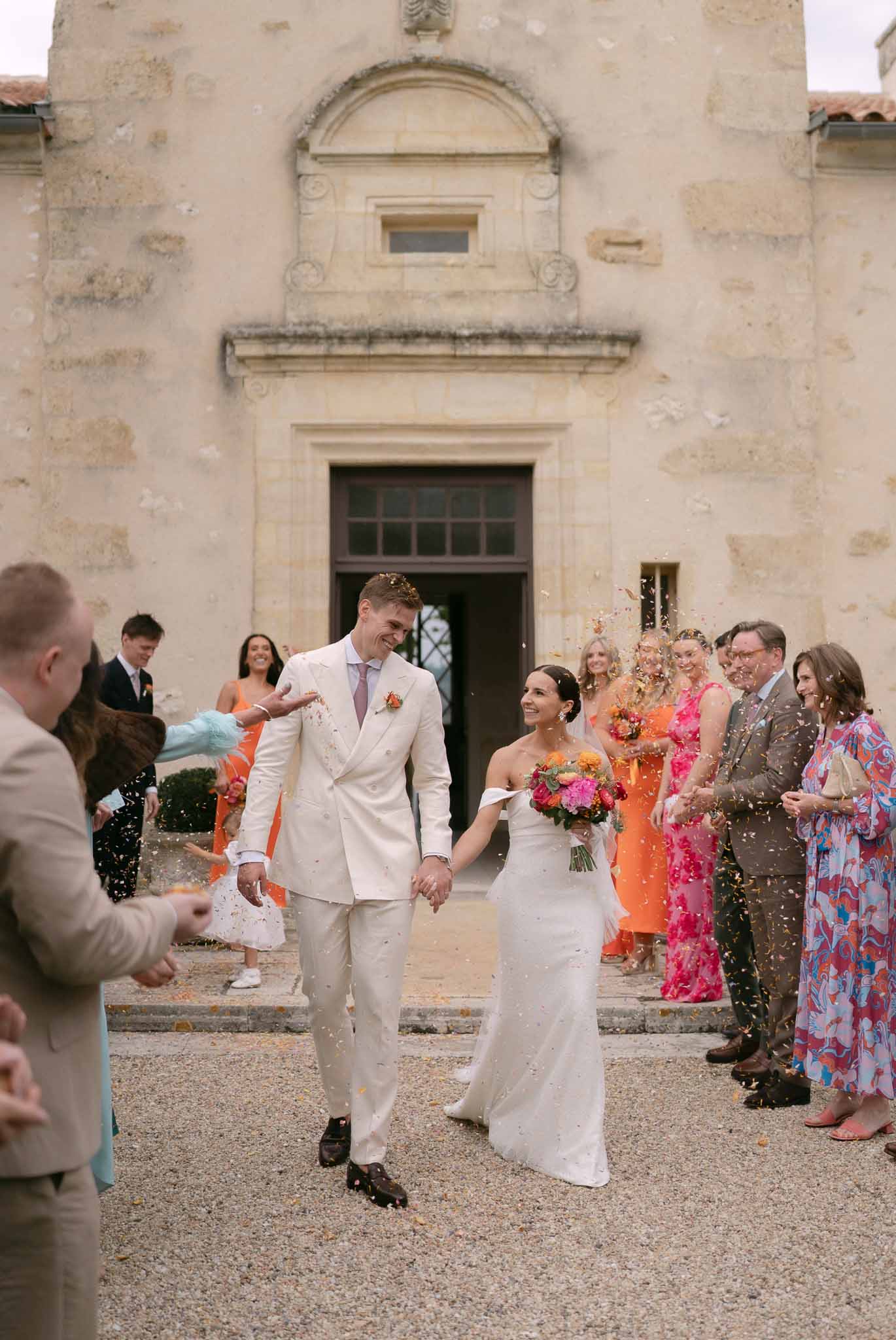 Bride and groom walk through confetti exit with guests throwing orange and pink petals outside limestone chateau