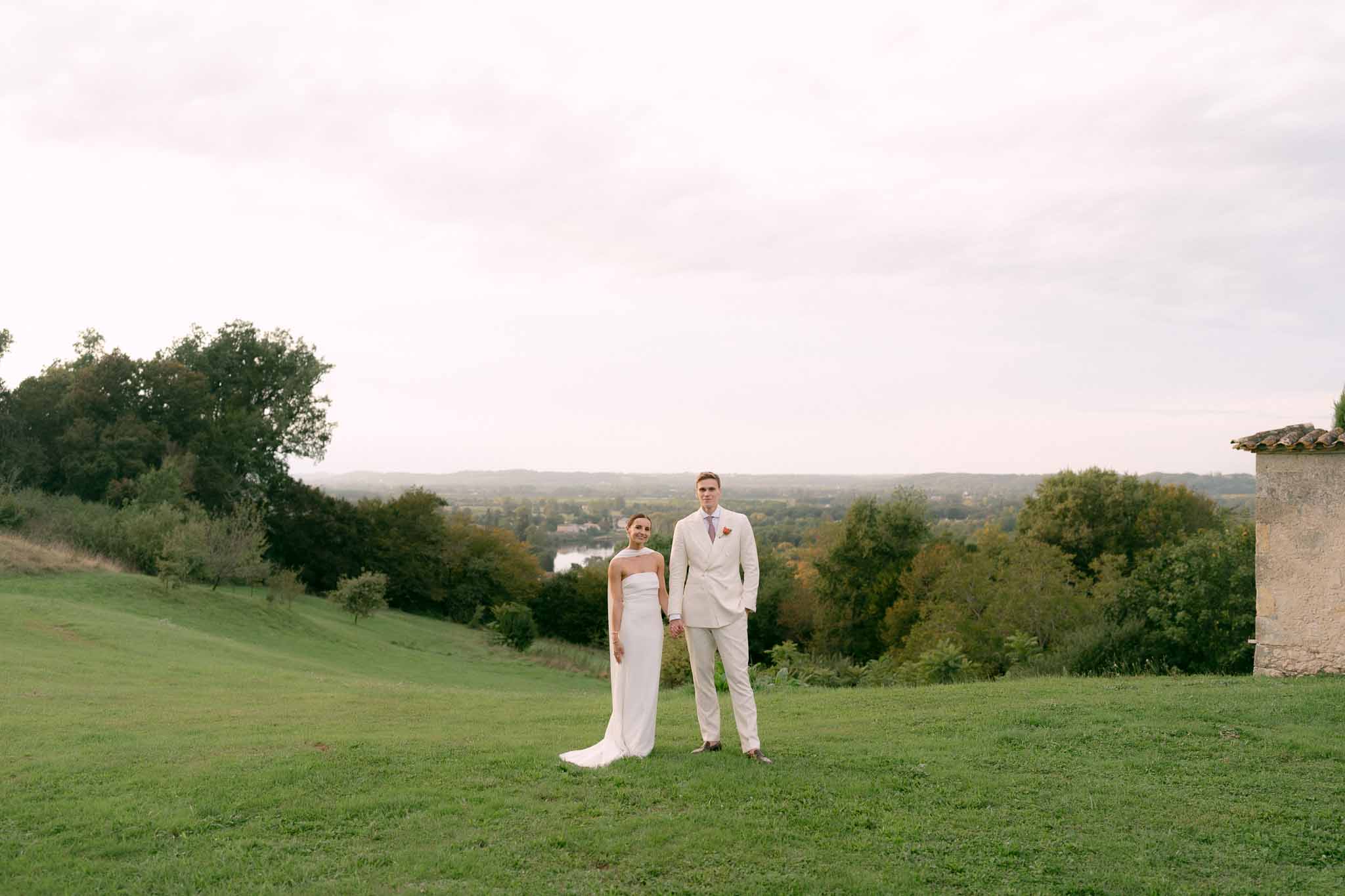 Full-length couple on chateau lawn holding hands bride in strapless gown groom in cream suit with countryside panorama