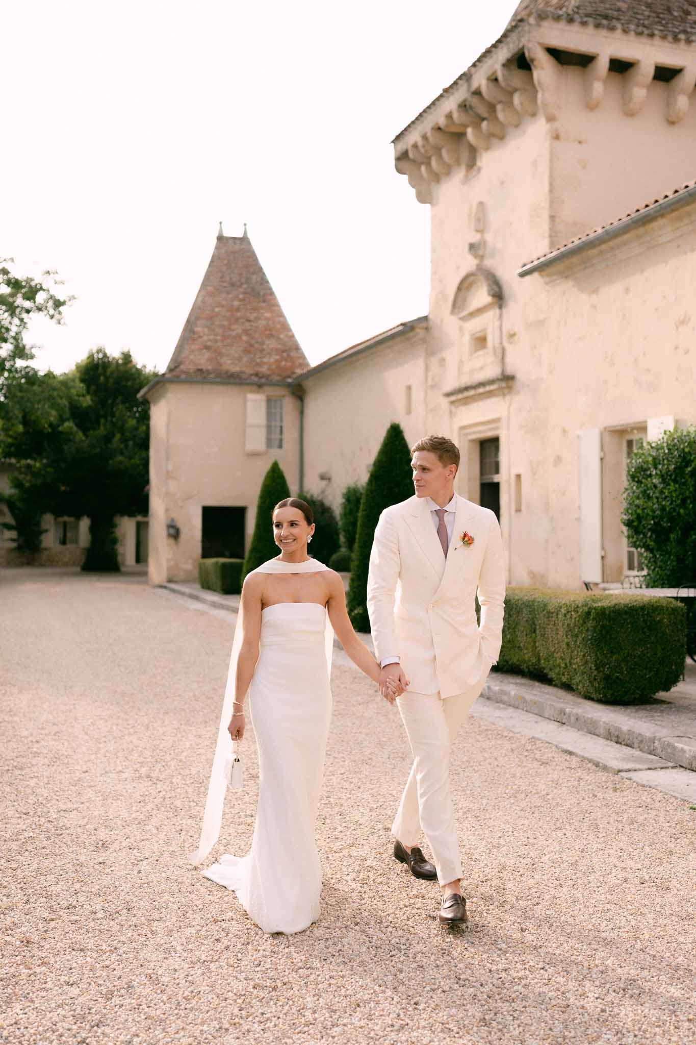 Couple walking hand in hand on chateau courtyard in golden hour light with topiary backdrop