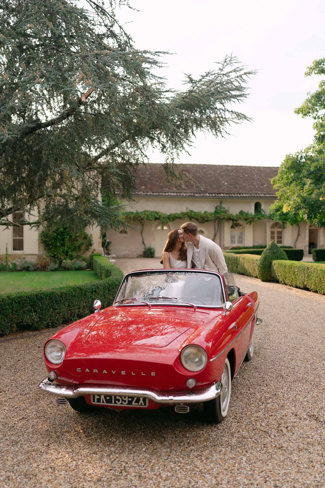 Bride and groom kissing behind vintage red Renault convertible on chateau gravel driveway