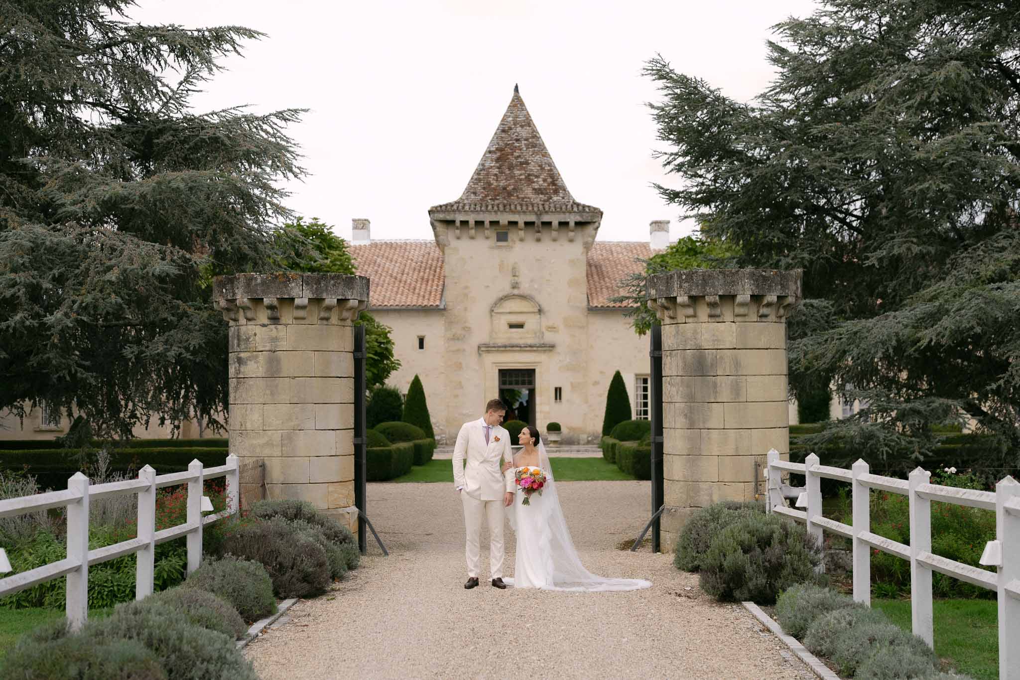 Couple between stone gate pillars of chateau with crenellated towers and vibrant orange bouquet