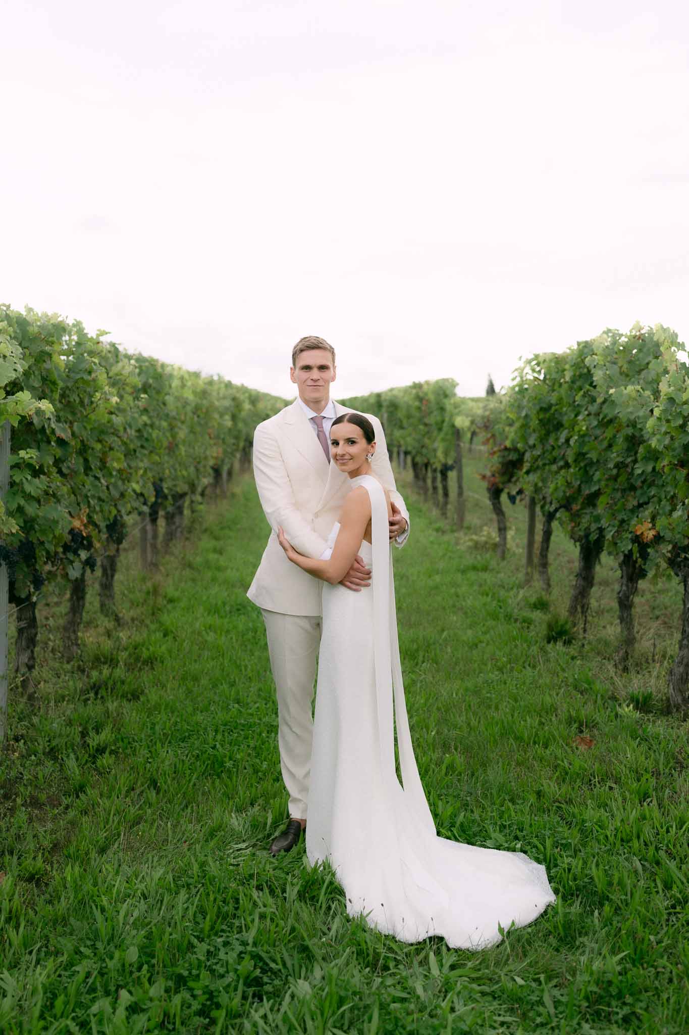 Bride in white gown with cape veil and groom in cream suit embracing between vineyard rows