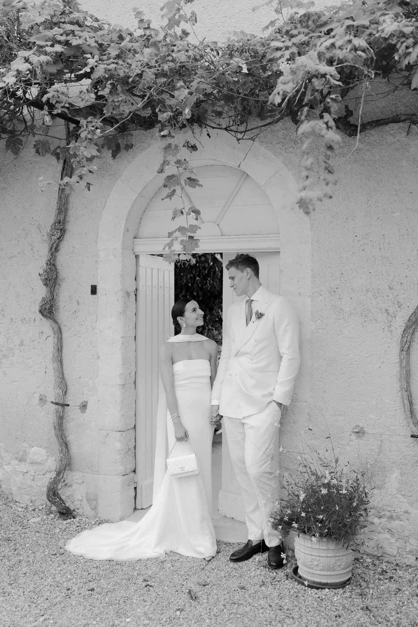 Black and white couple holding hands before vine-framed arched doorway, bride in halter gown with clutch