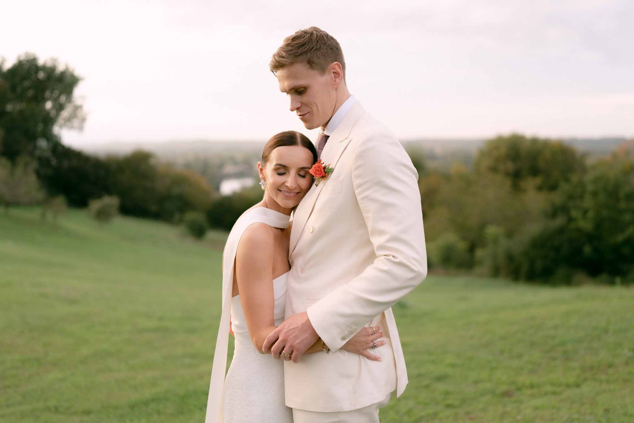 Bride in white halter dress leaning into groom in cream double-breasted suit with coral boutonniere outdoors
