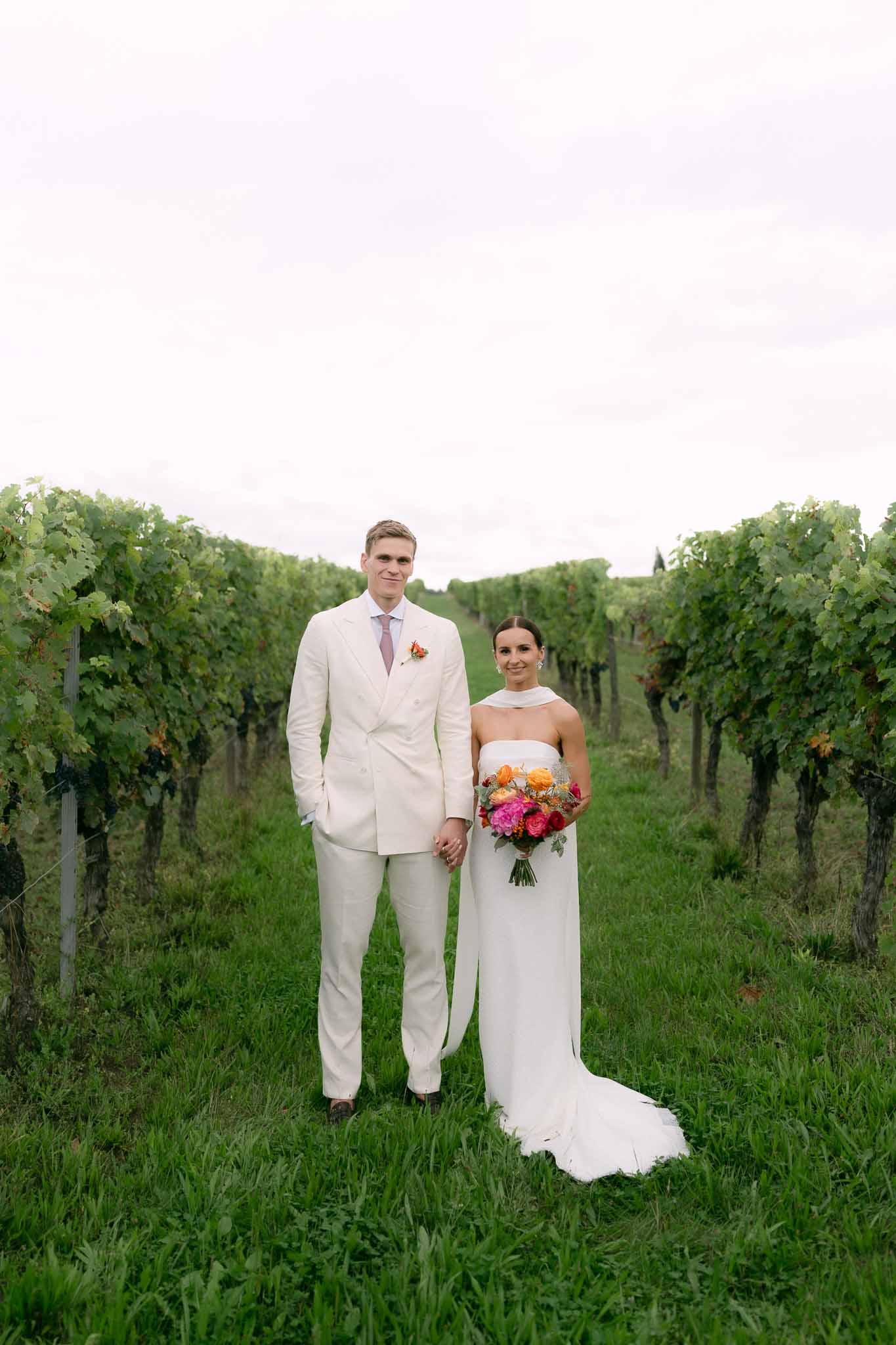Bride and groom hand in hand in vineyard, bride holding hot pink and orange bouquet, groom in cream suit