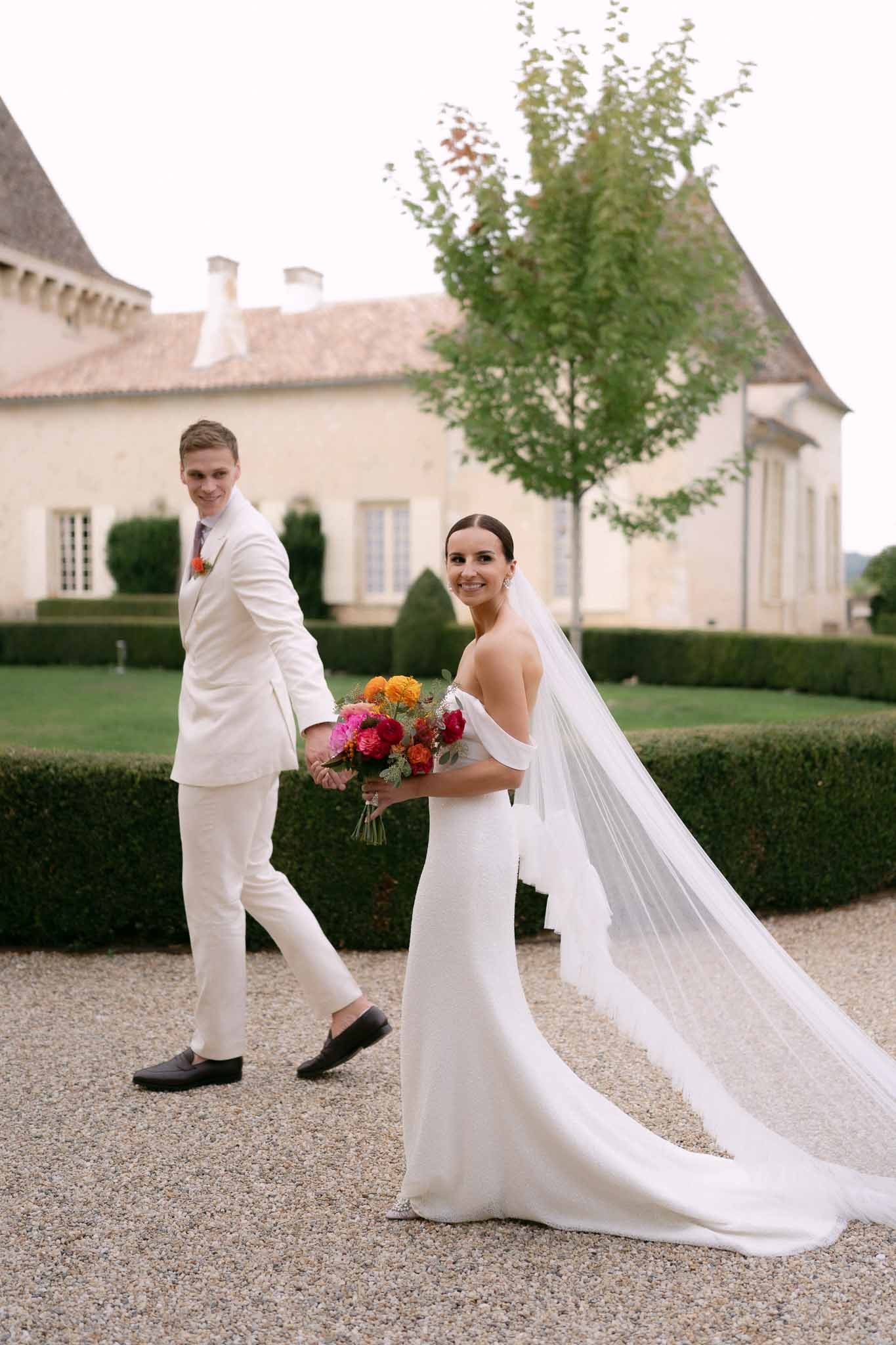 Groom in ivory suit leads bride with cathedral veil and vibrant pink and orange bouquet on chateau gravel path