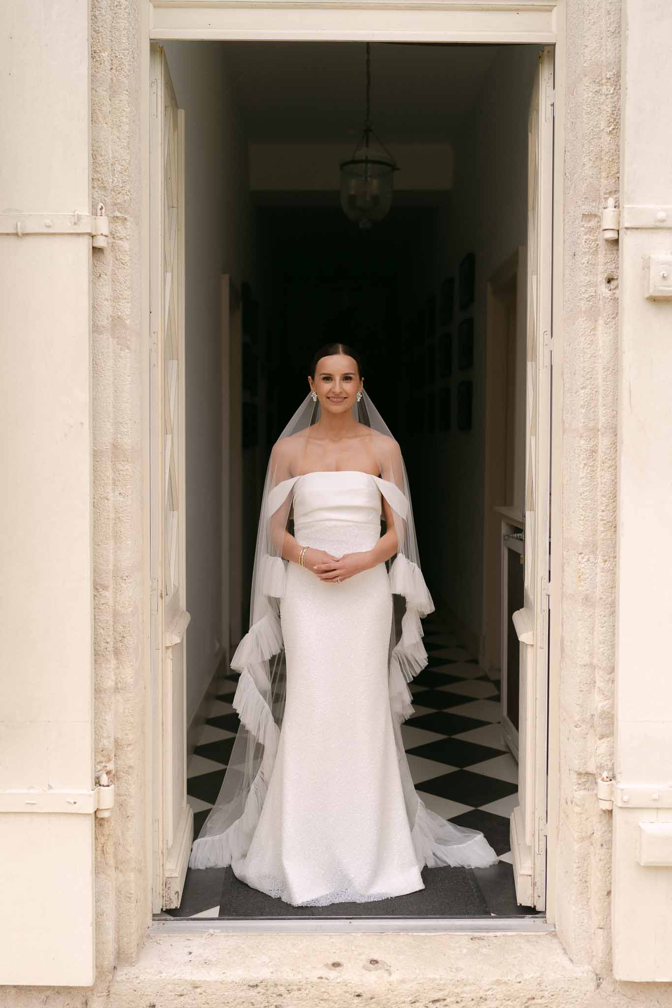 Bride in strapless column gown with ruffled tulle cathedral veil framed by stone doorway and diamond-tile hallway