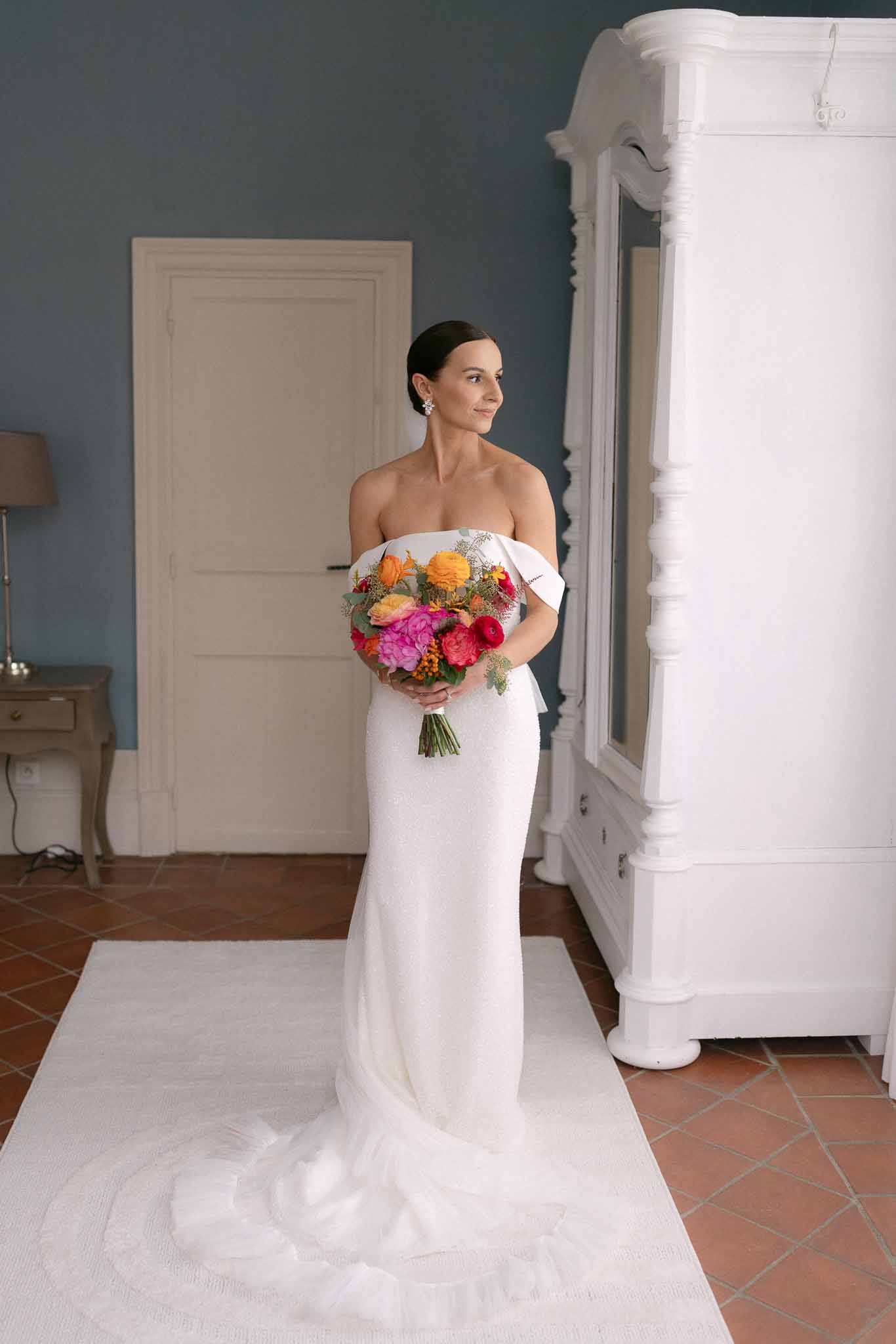 Bride in off-shoulder white gown holding vibrant bouquet of pink peonies, orange dahlias, and coral roses in chateau room