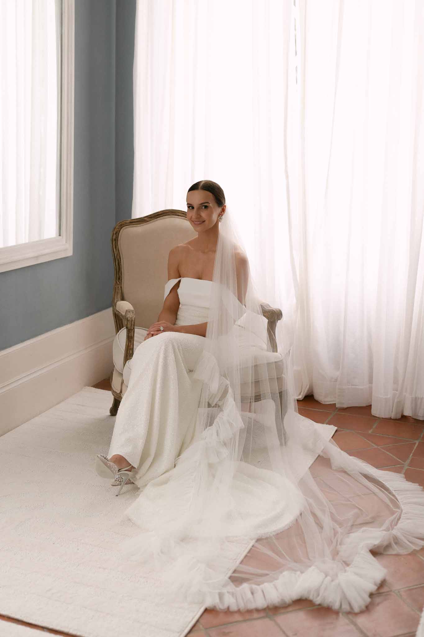 Bride in ivory strapless column gown with cathedral veil seated in Louis armchair in blue-grey room