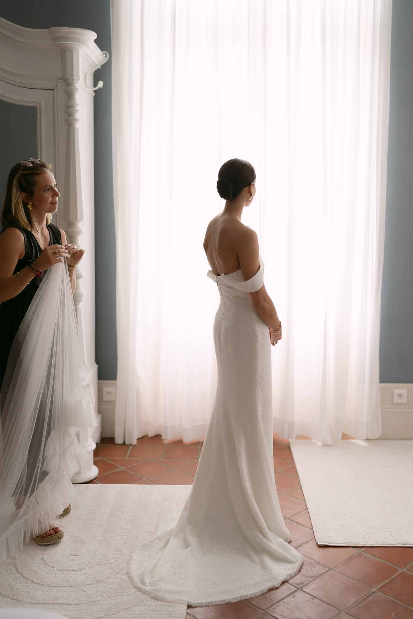Bride in off-shoulder gown with bow detail seen from behind as attendant holds ruffled tulle veil