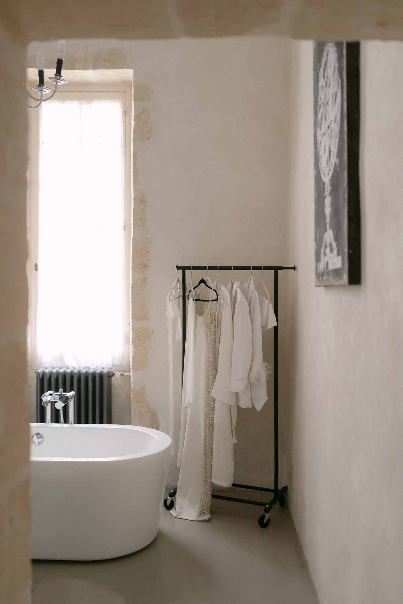 Garment rail with wedding dress and robes in minimalist bathroom with freestanding bathtub