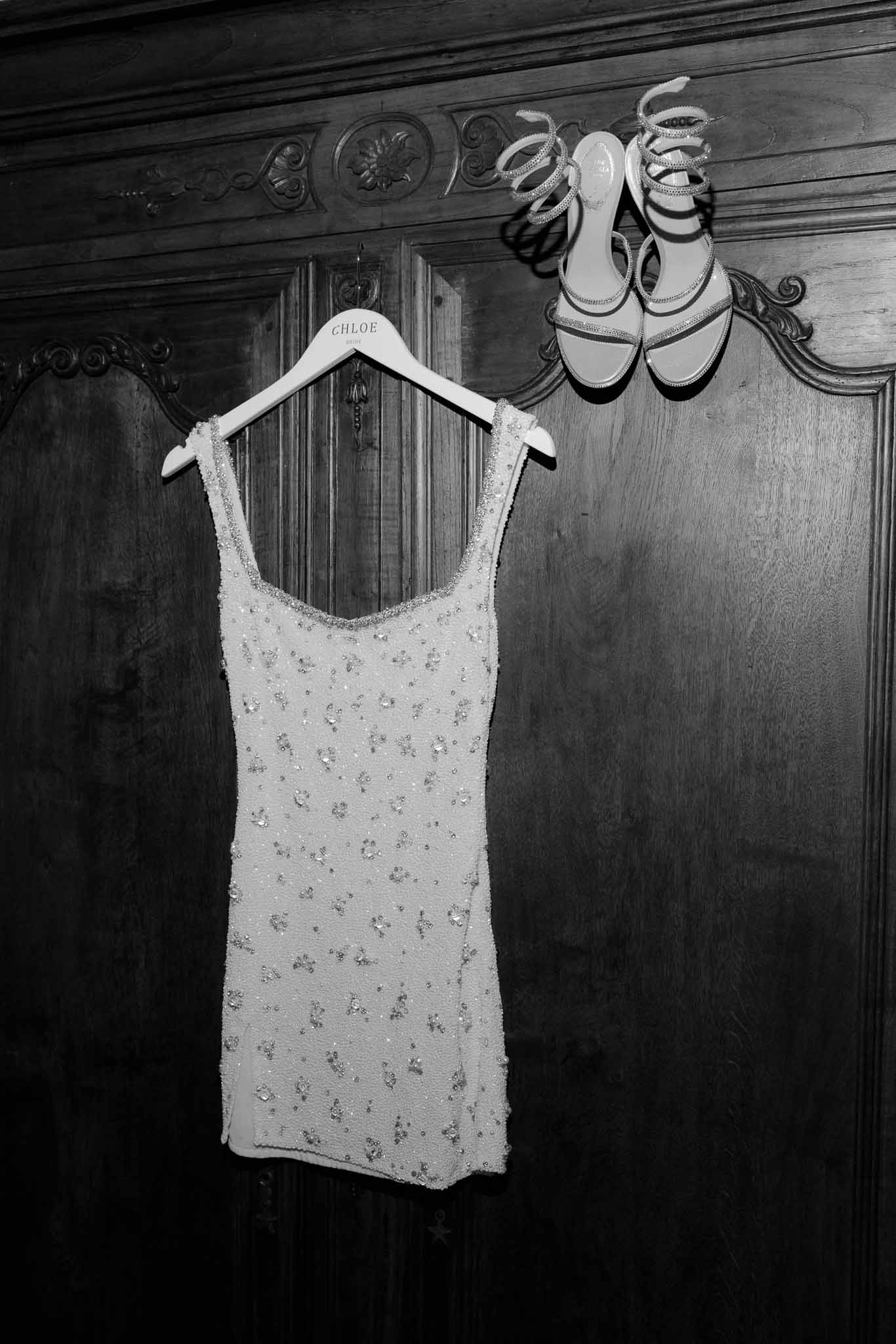 Embellished mini bridal dress and crystal sandals hanging on antique armoire in black and white