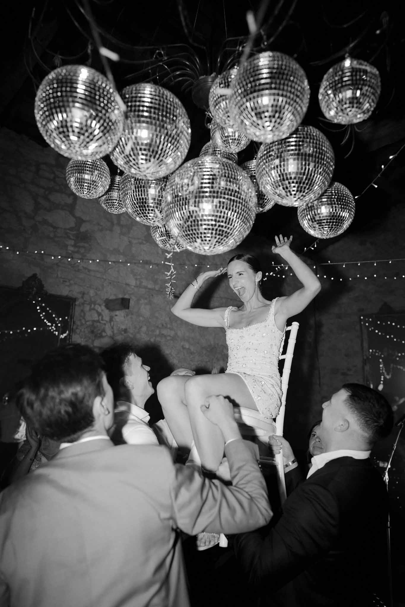 Black and white low-angle shot of bride lifted on chair beneath cluster of mirror disco balls