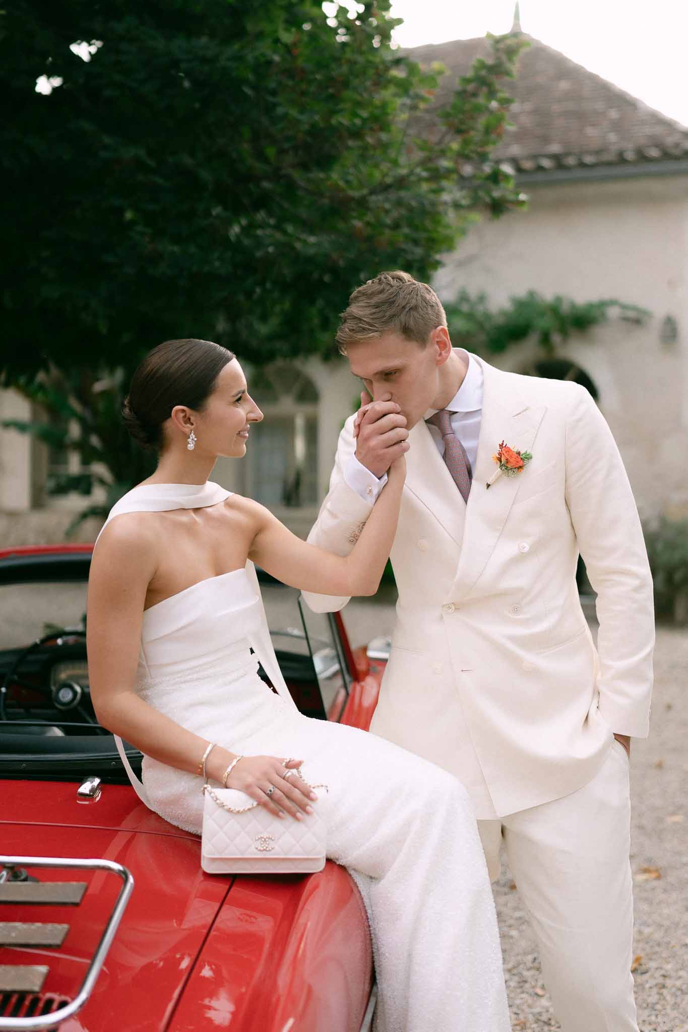 Groom in cream suit kissing bride's hand as she sits on a vintage red convertible at a French estate