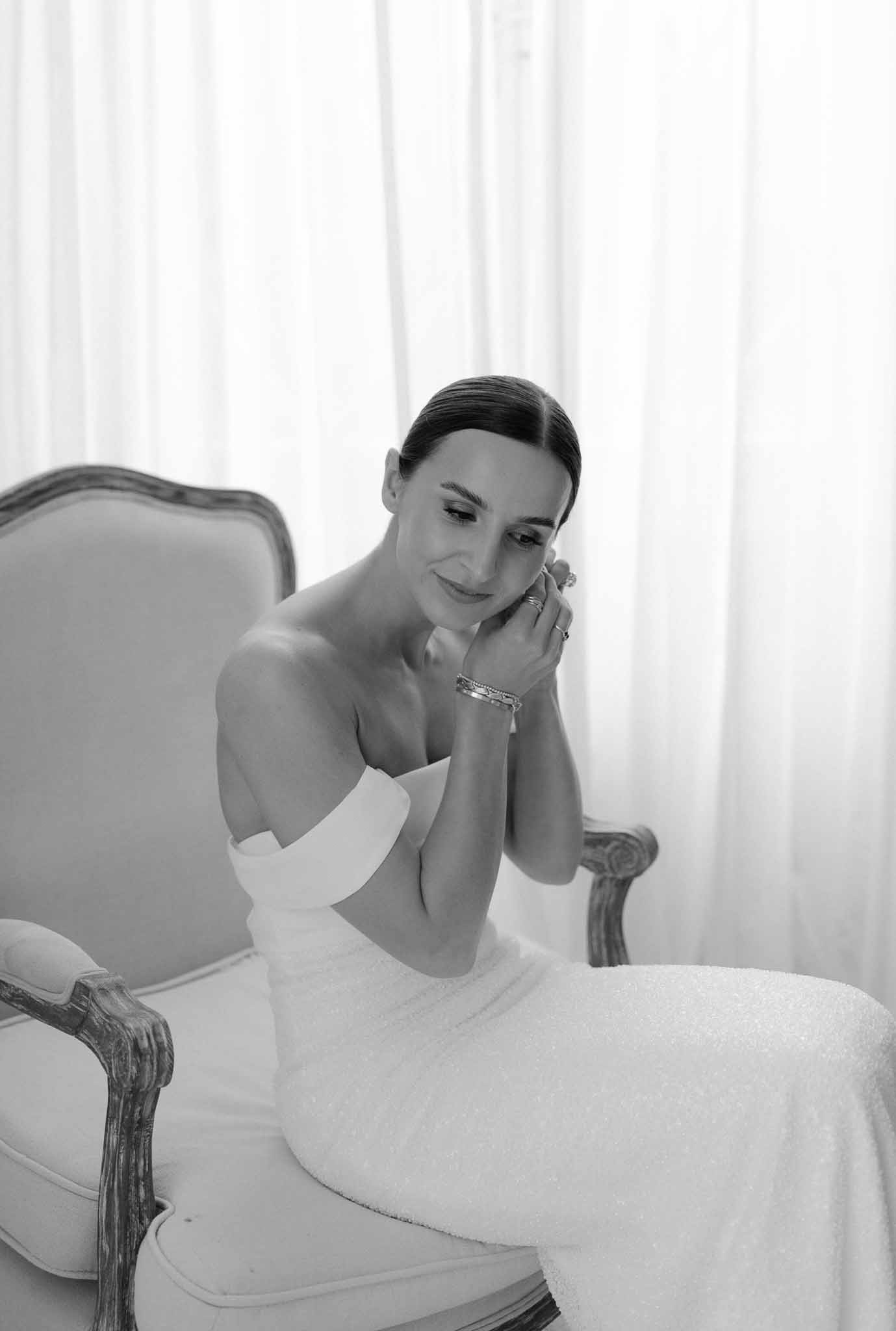 Black and white portrait of bride in embellished strapless gown seated in Louis armchair by sheer curtains