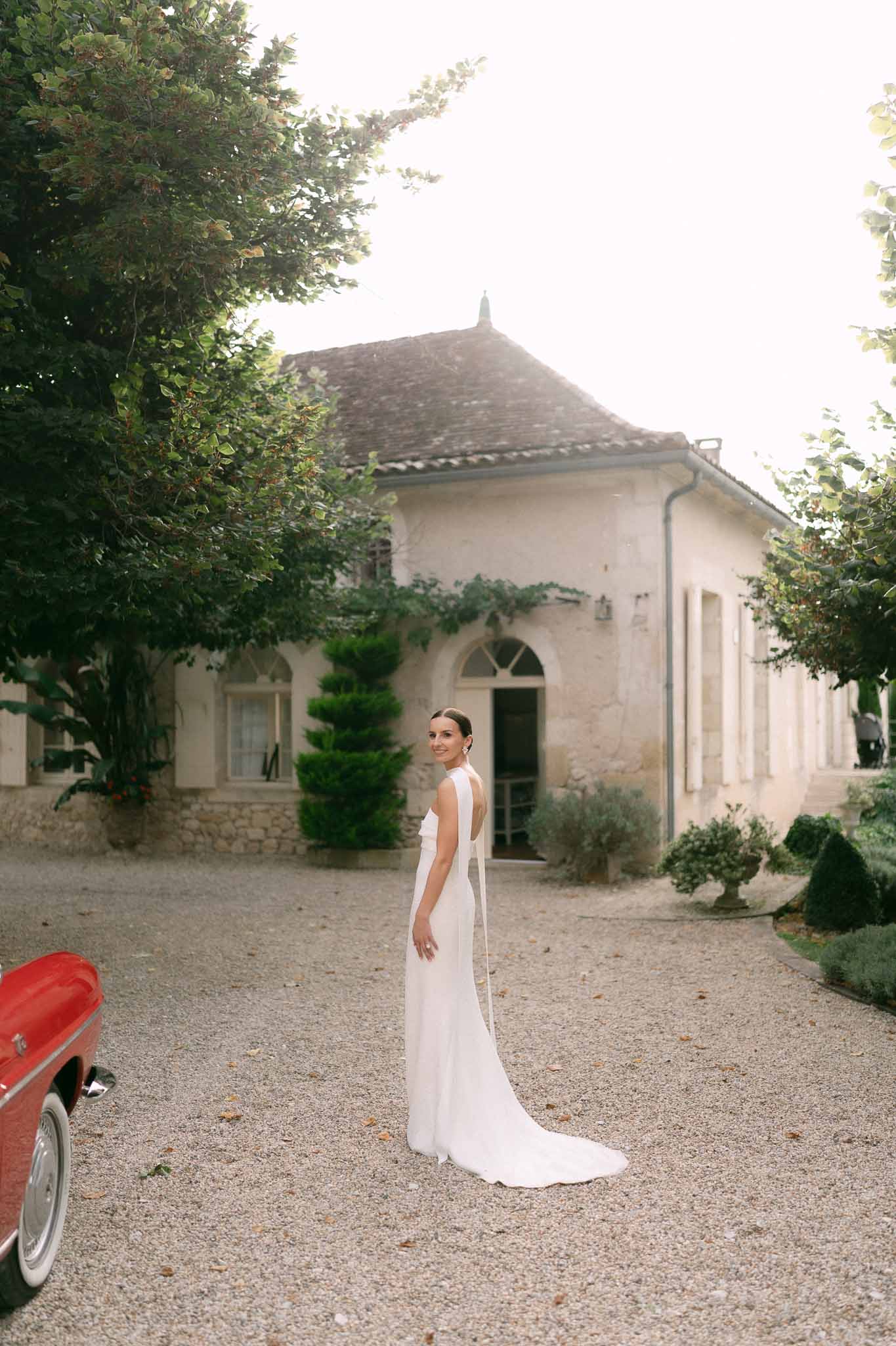 Bride in halter-neck open-back gown glances over shoulder beside vintage red convertible at stone estate