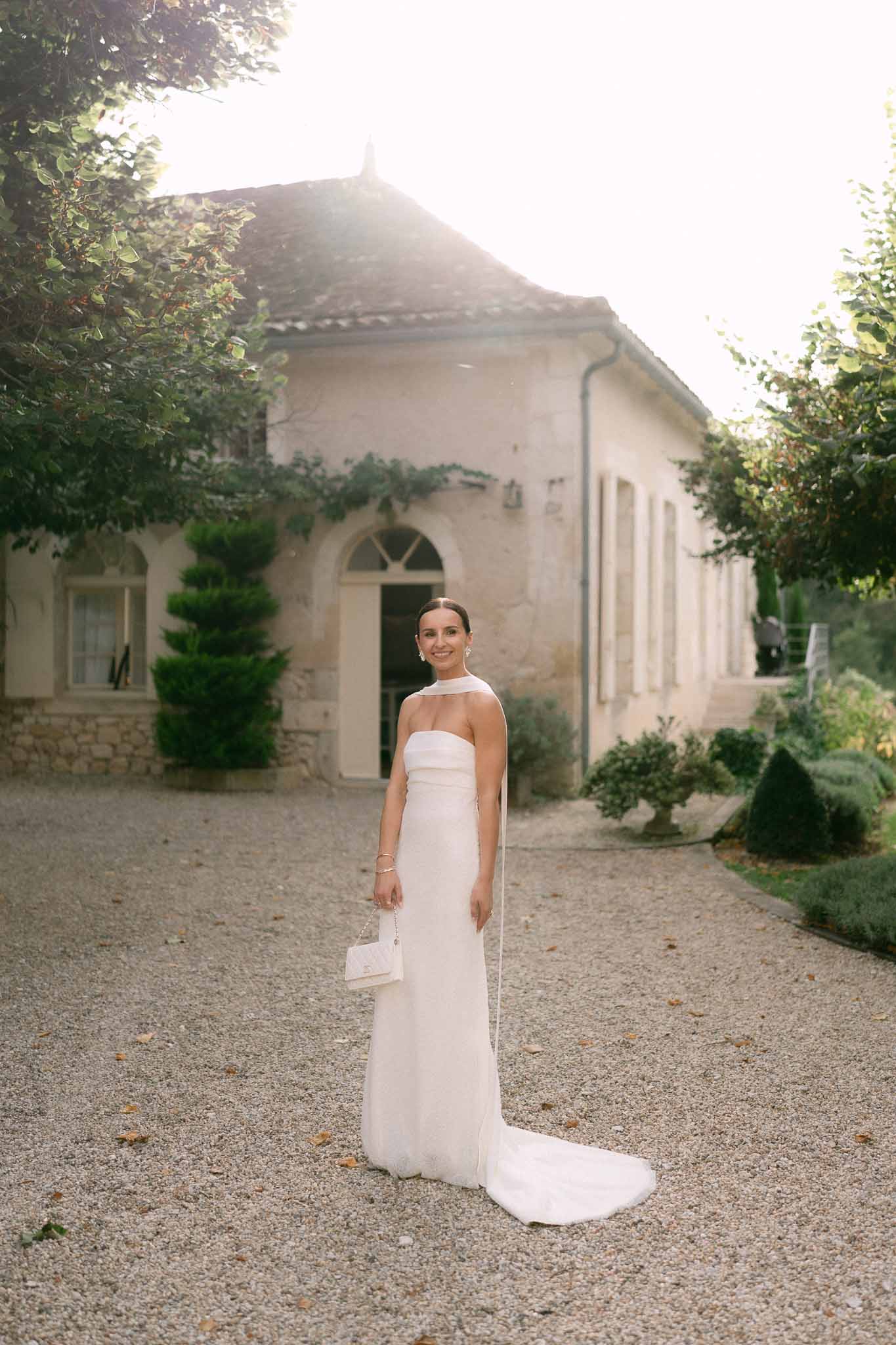 Bride in white halter gown with trailing scarf standing on gravel courtyard before French stone country house