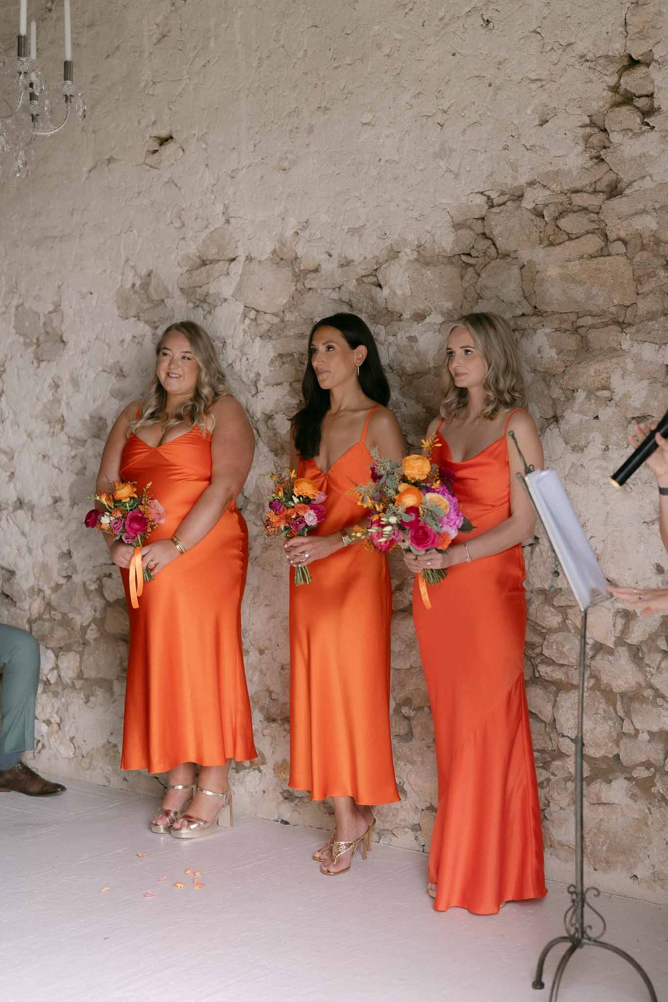 Three bridesmaids in burnt orange satin dresses holding vibrant pink and orange bouquets during a stone-walled ceremony