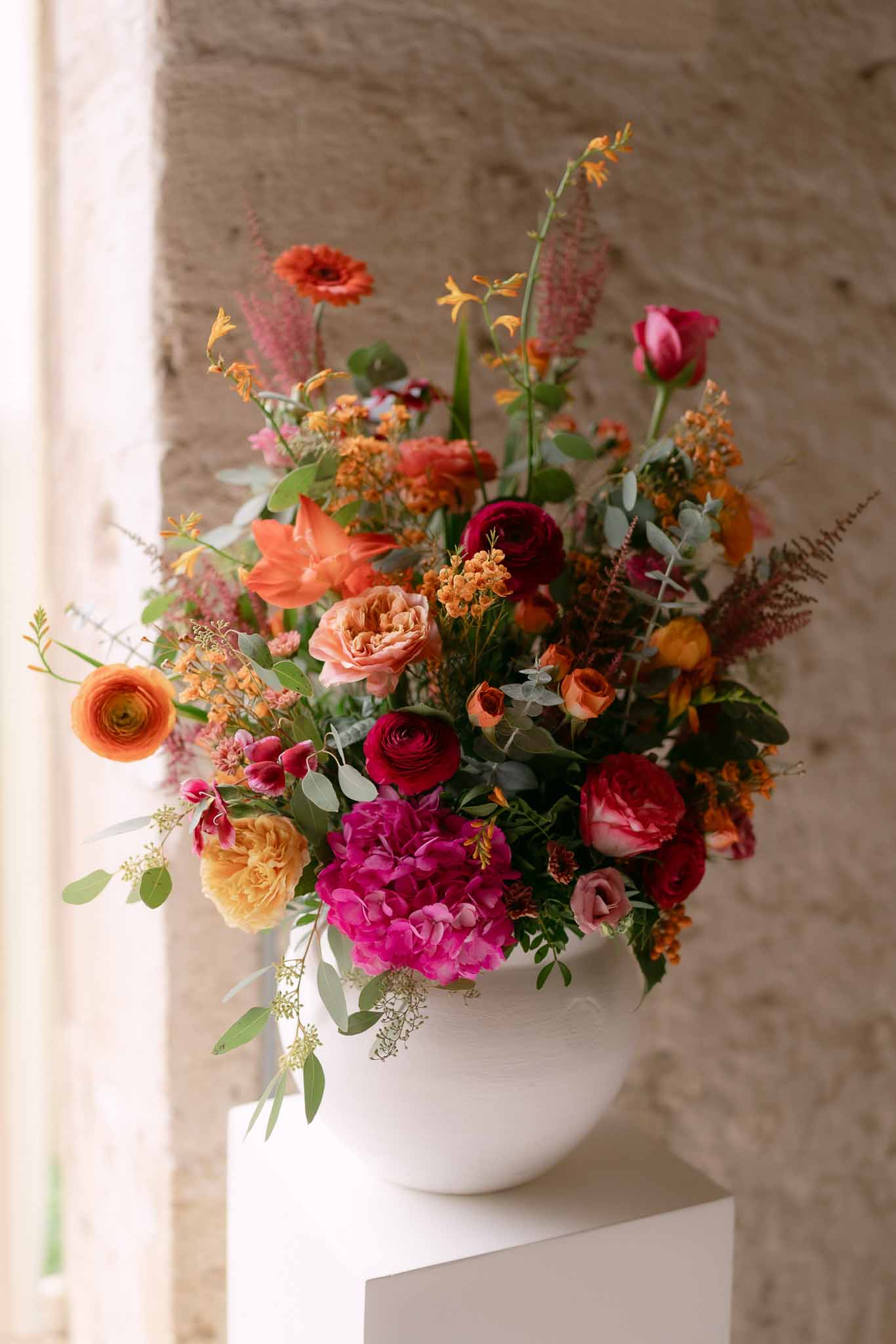 Magenta peonies, burgundy ranunculus, and orange roses in white ceramic urn against stone wall