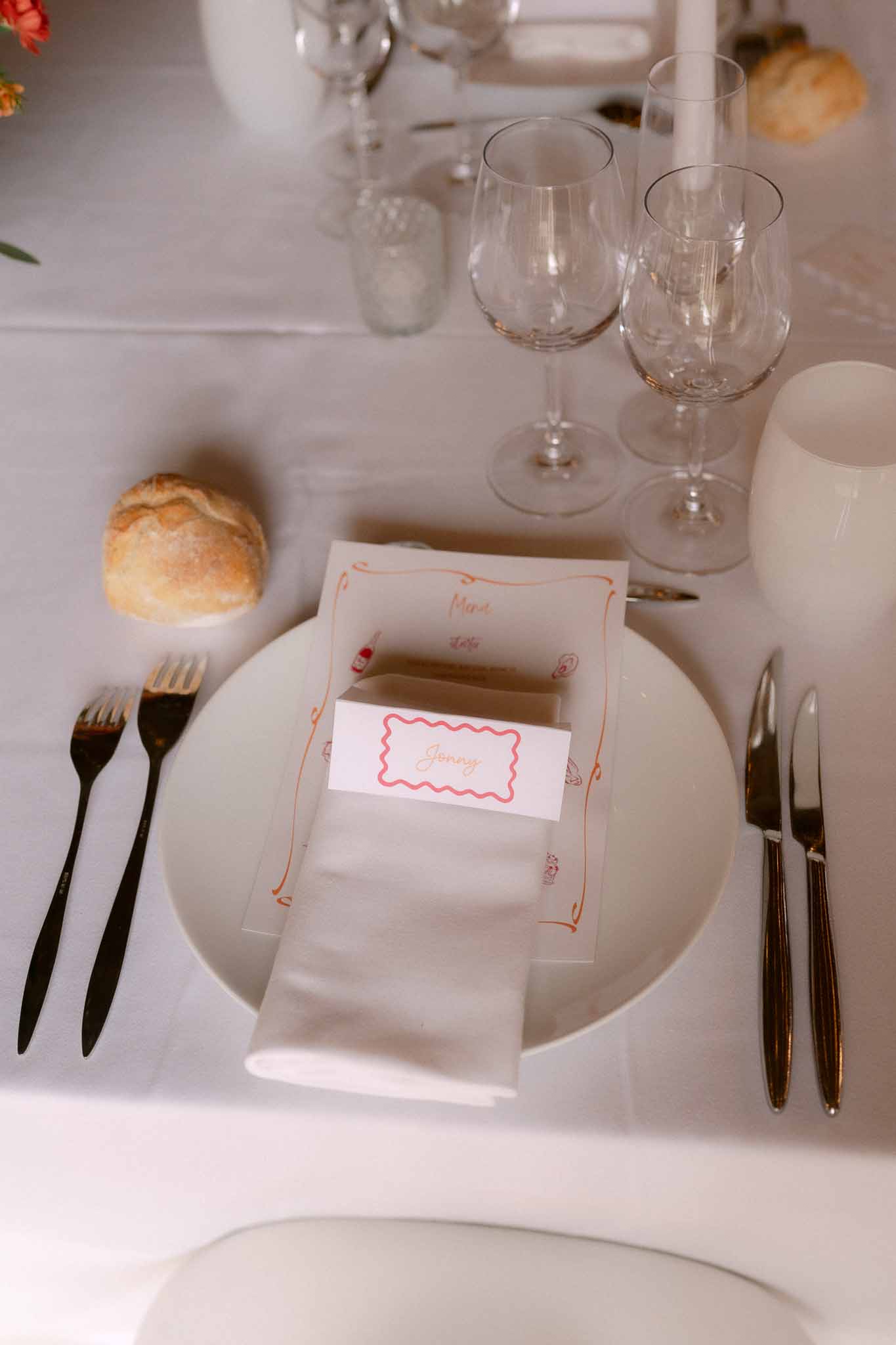Wedding place setting with white plate, printed menu card with orange border, place card, black matte cutlery, and crystal...