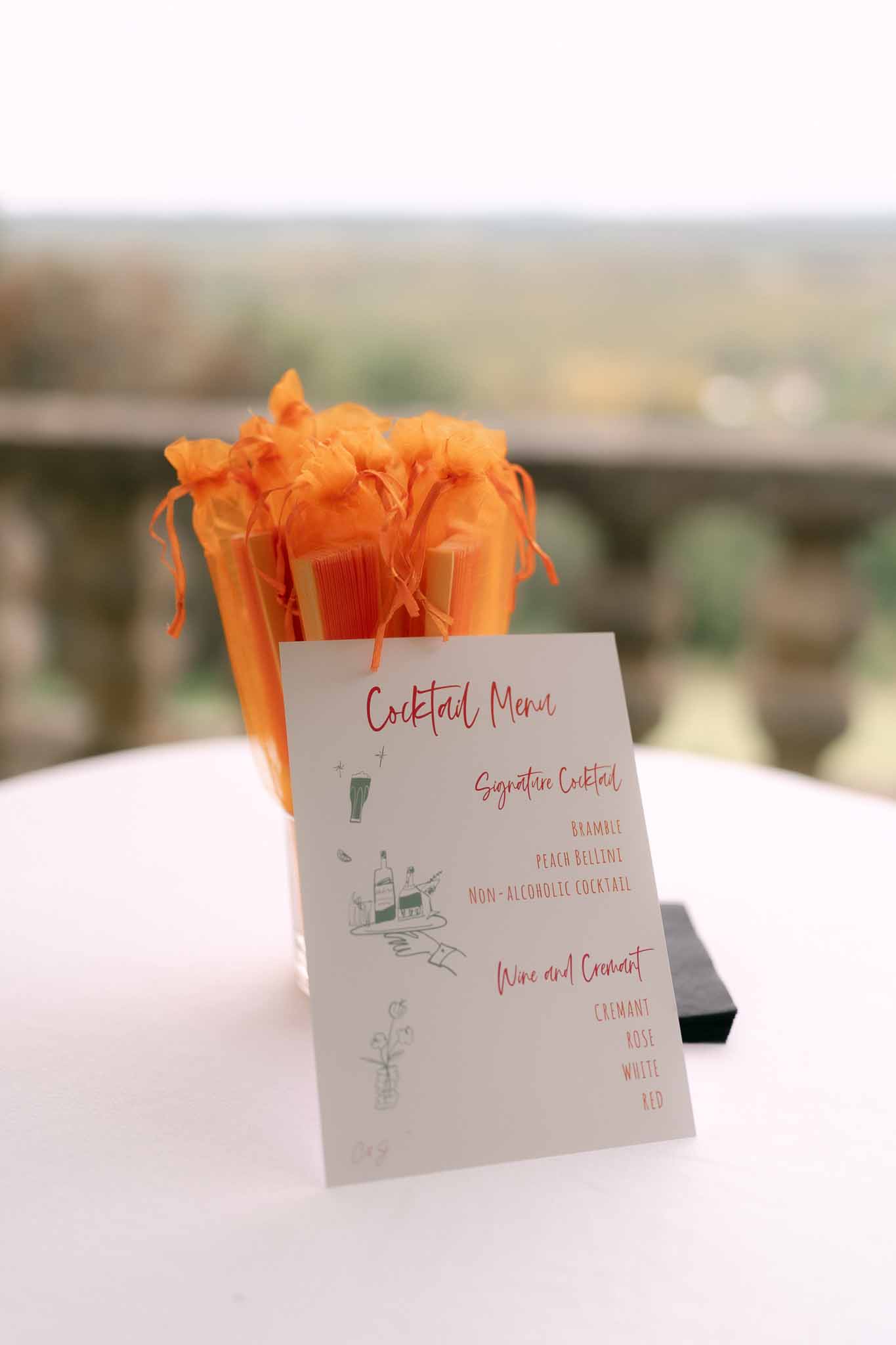 White cocktail menu card listing signature drinks beside orange organza favor bags on terrace table