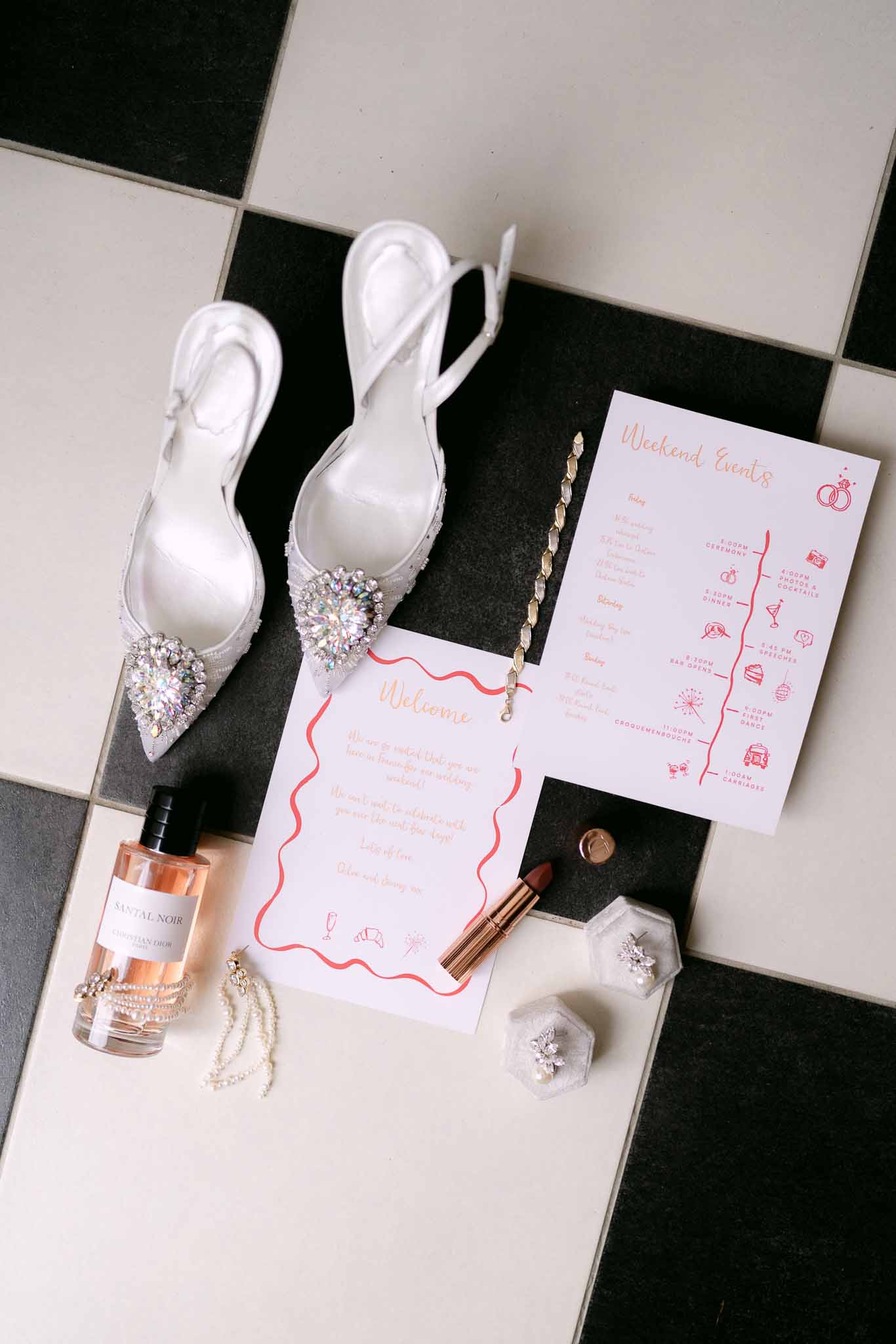Flat lay of bridal accessories including crystal heels, Dior perfume, pearl earrings, and coral stationery