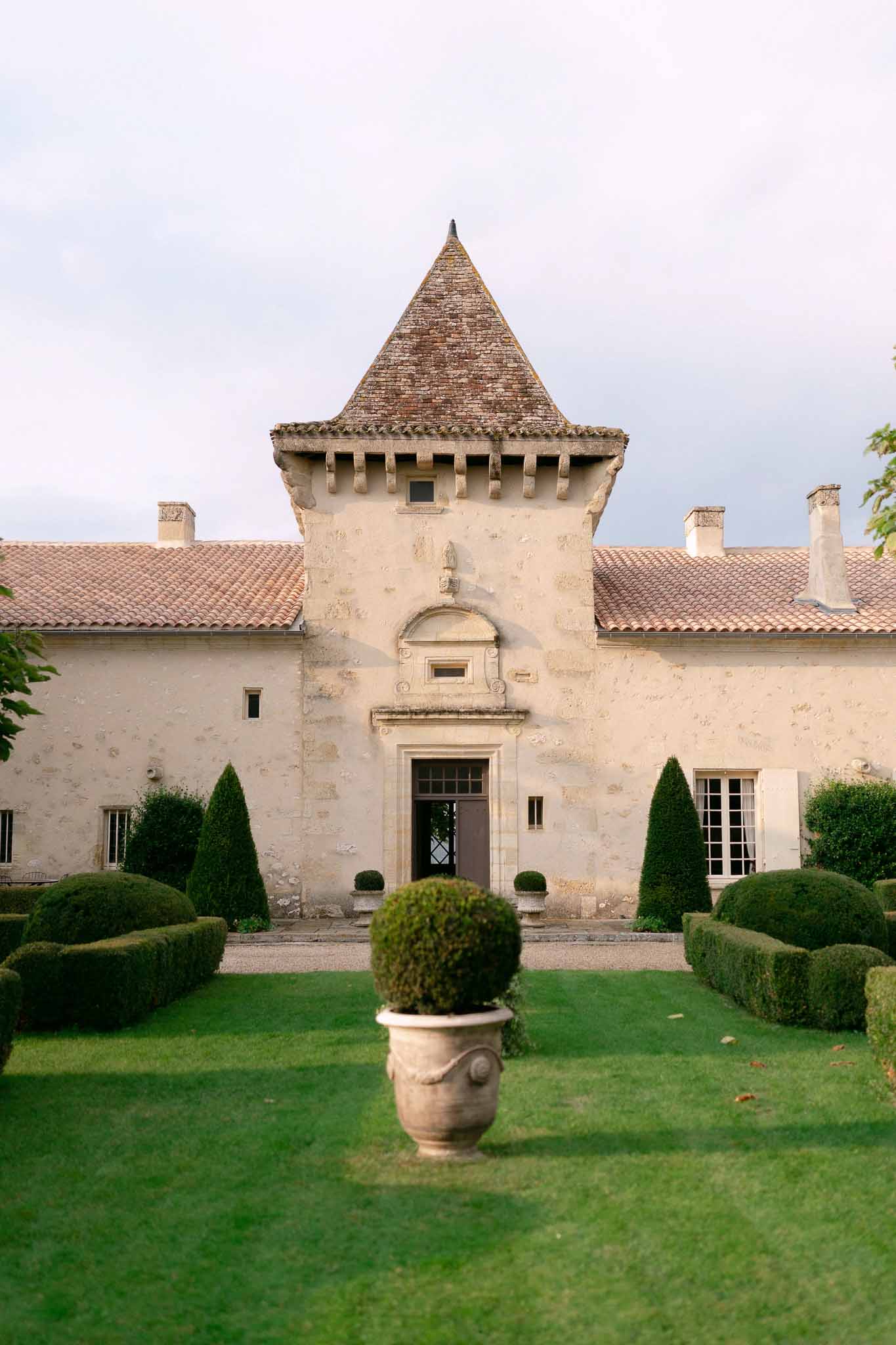Chateau facade with central stone tower, conical roof, symmetrical topiary, and clipped box hedging