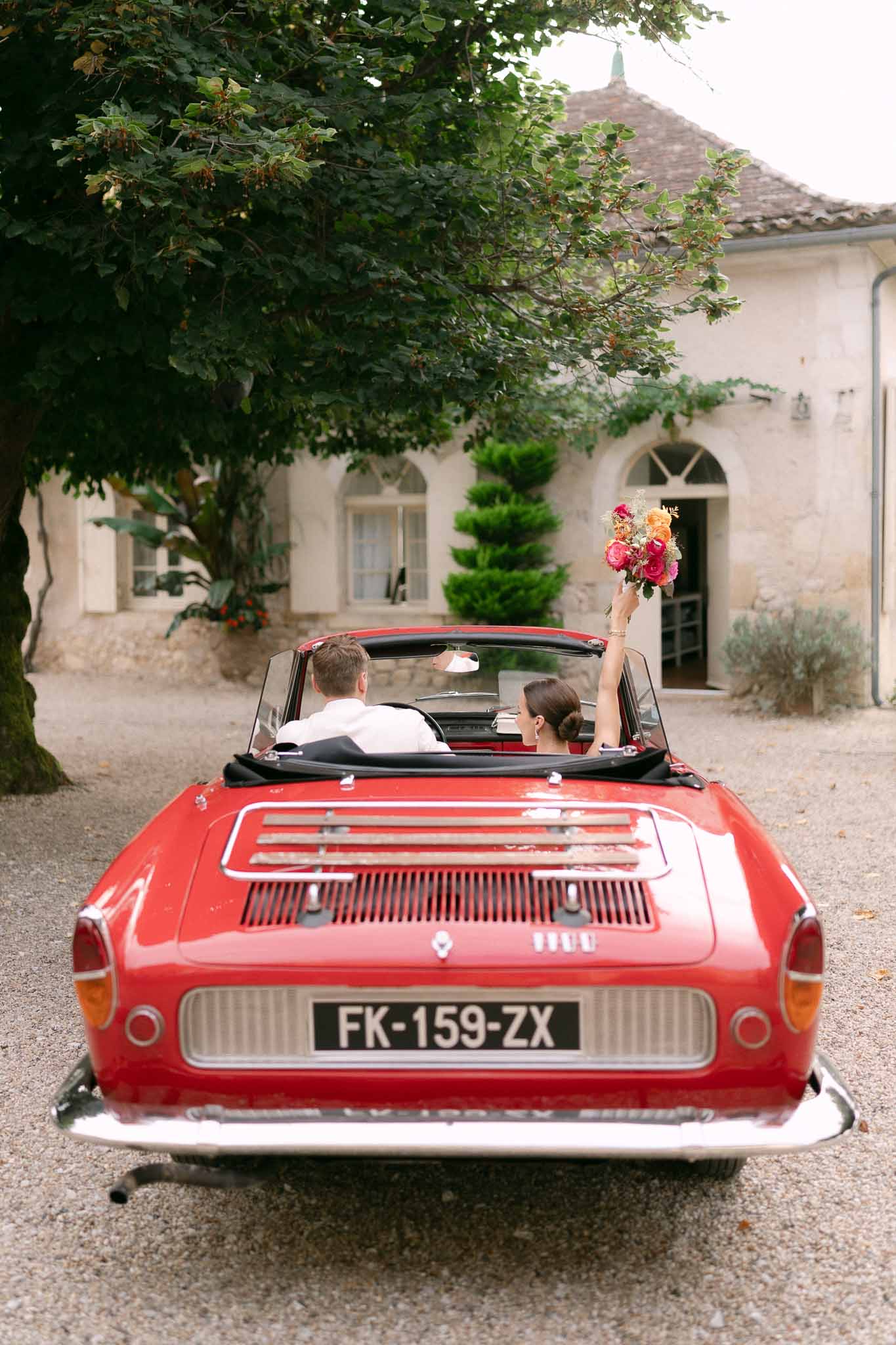 Bride raising colorful bouquet from a vintage red Renault convertible on a chateau gravel driveway seen from behind