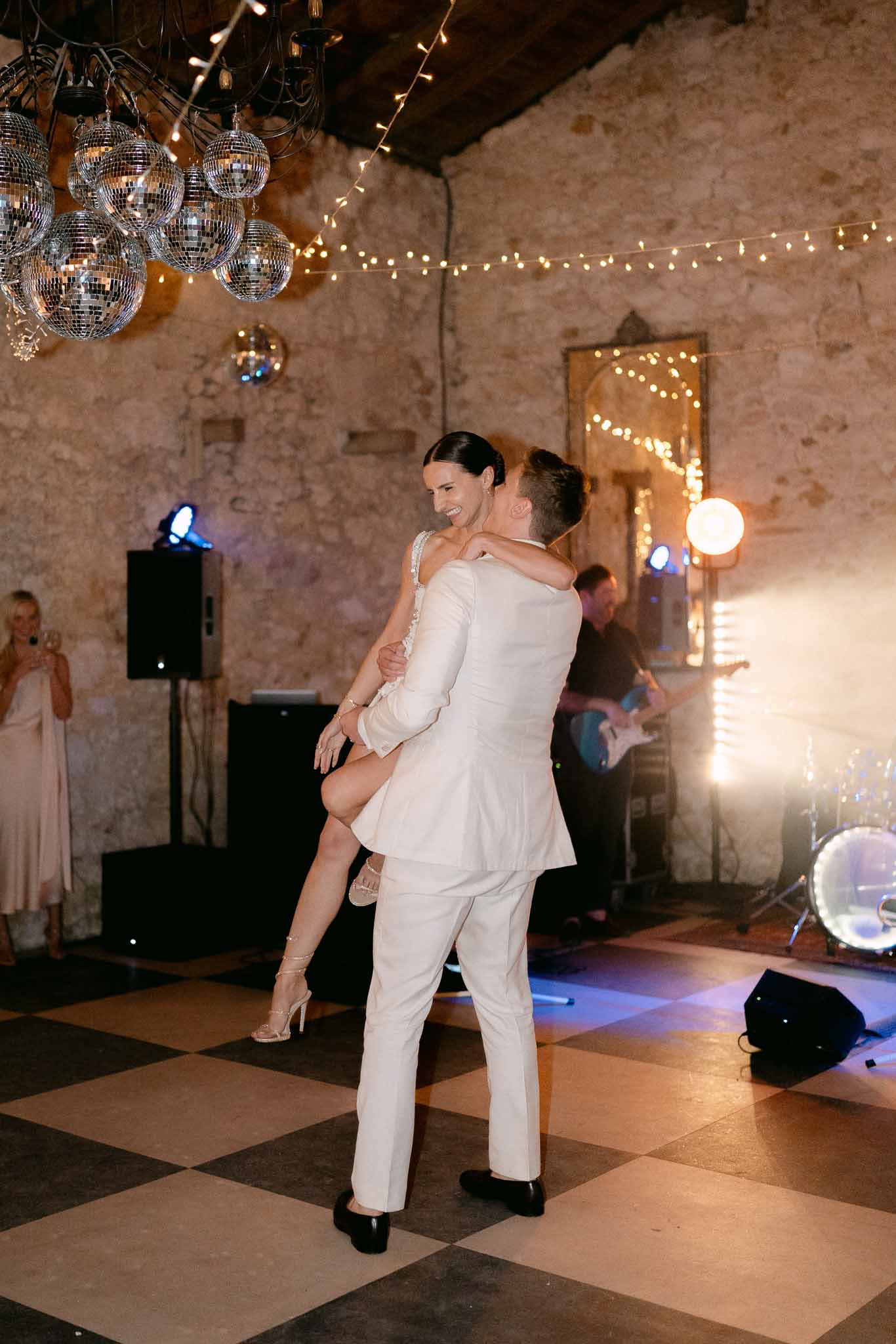 Groom lifts bride during first dance on checkerboard floor under disco balls and fairy lights with live band