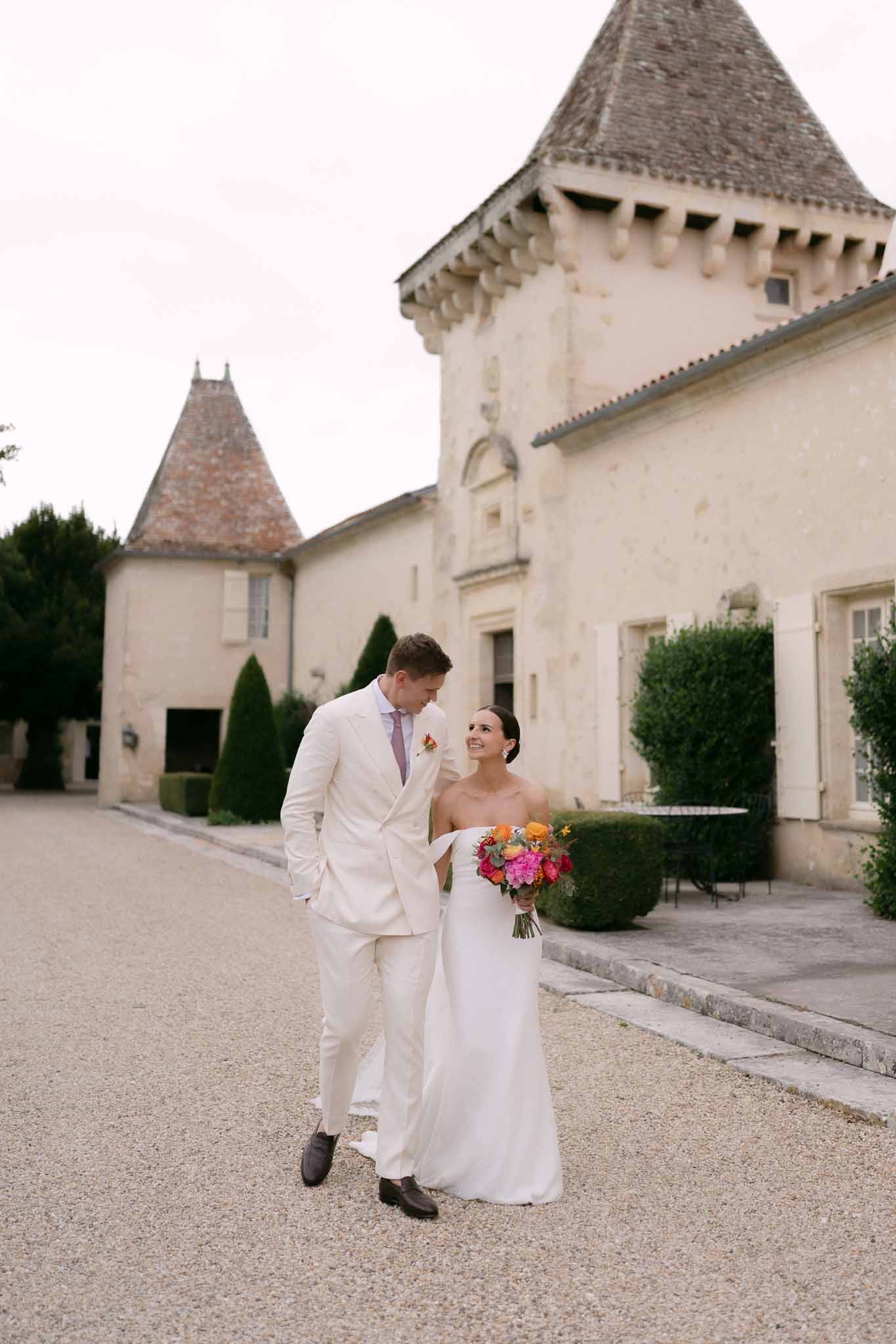 Couple walking on chateau gravel forecourt, bride carrying hot pink and orange bouquet