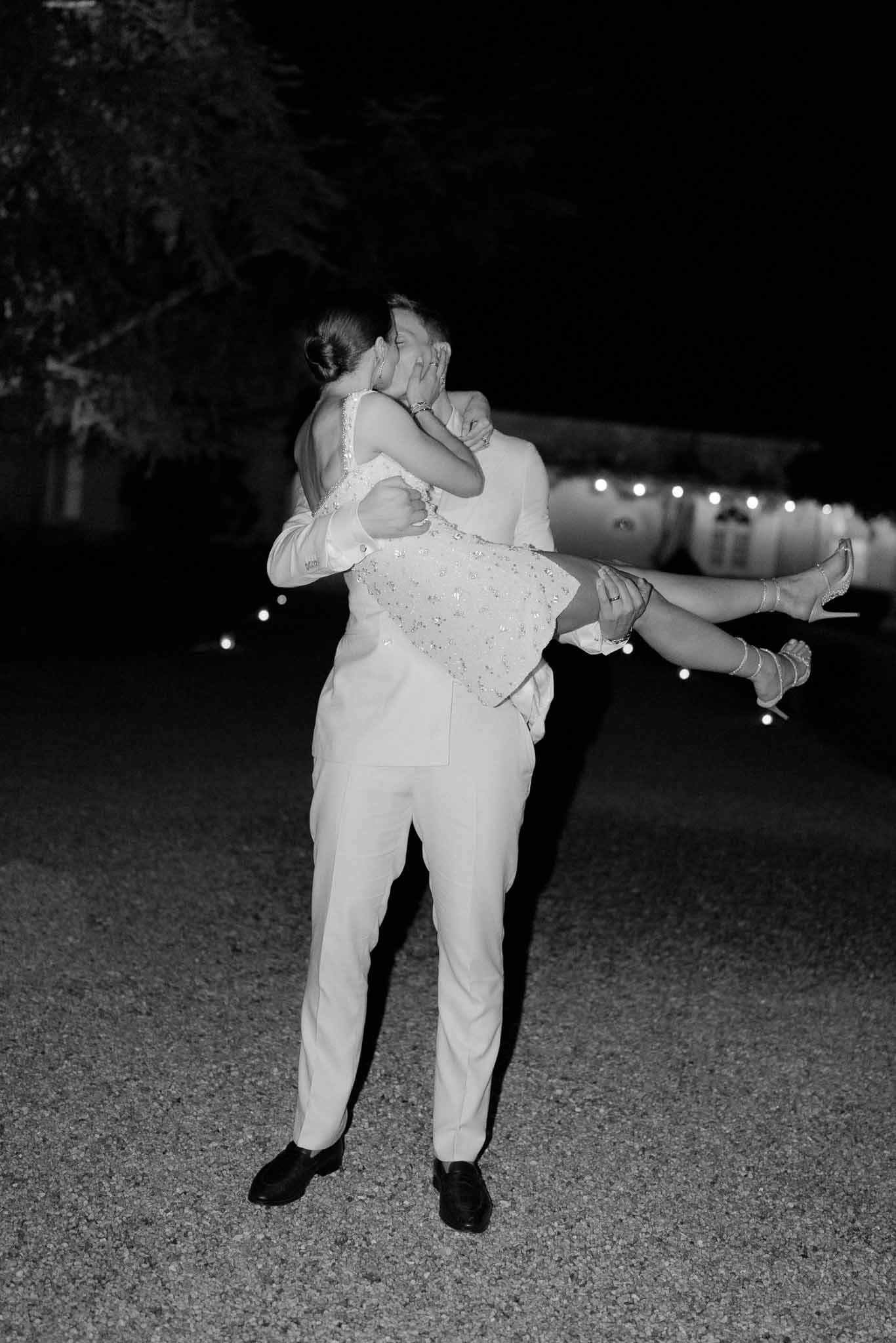 Black and white nighttime photo of groom lifting bride as they kiss on gravel with string lights behind
