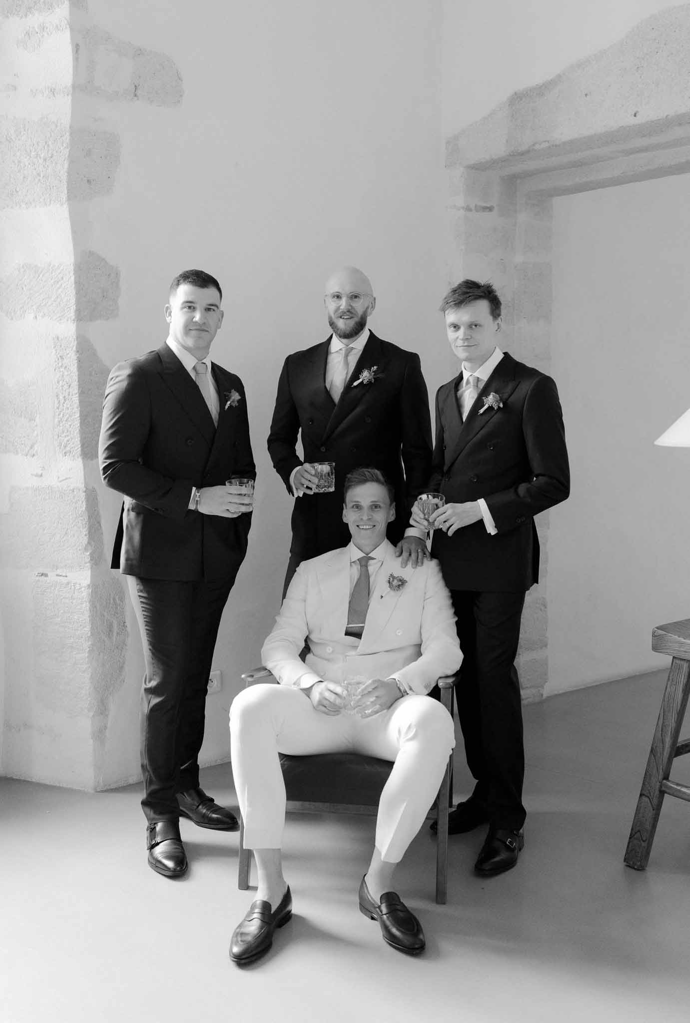 Black and white portrait of groom in light suit seated with three groomsmen in dark suits holding whisky