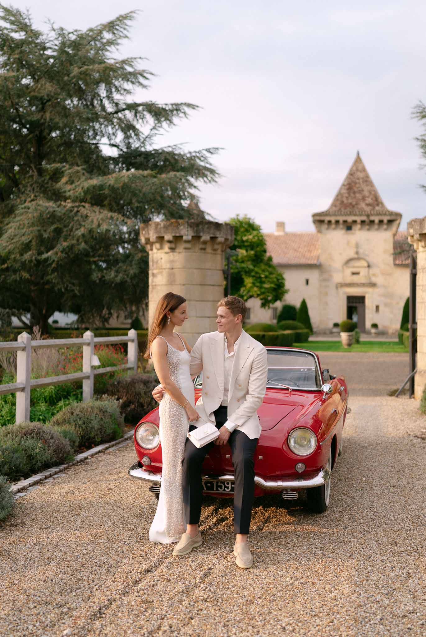 Couple poses beside red vintage convertible on gravel drive with turreted chateau behind