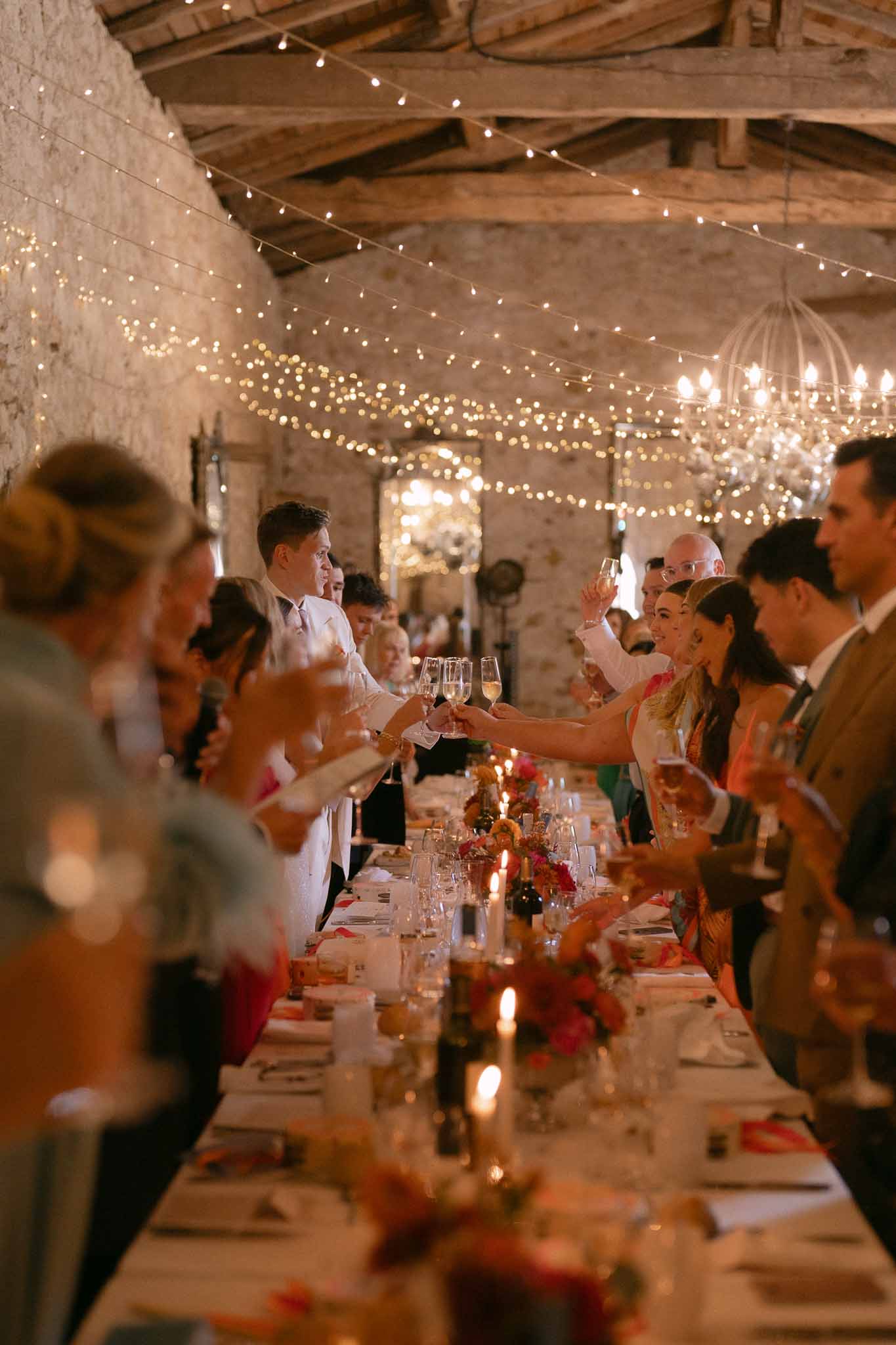 Groom in white suit toasts under fairy-lit barn ceiling with red and orange floral feasting table
