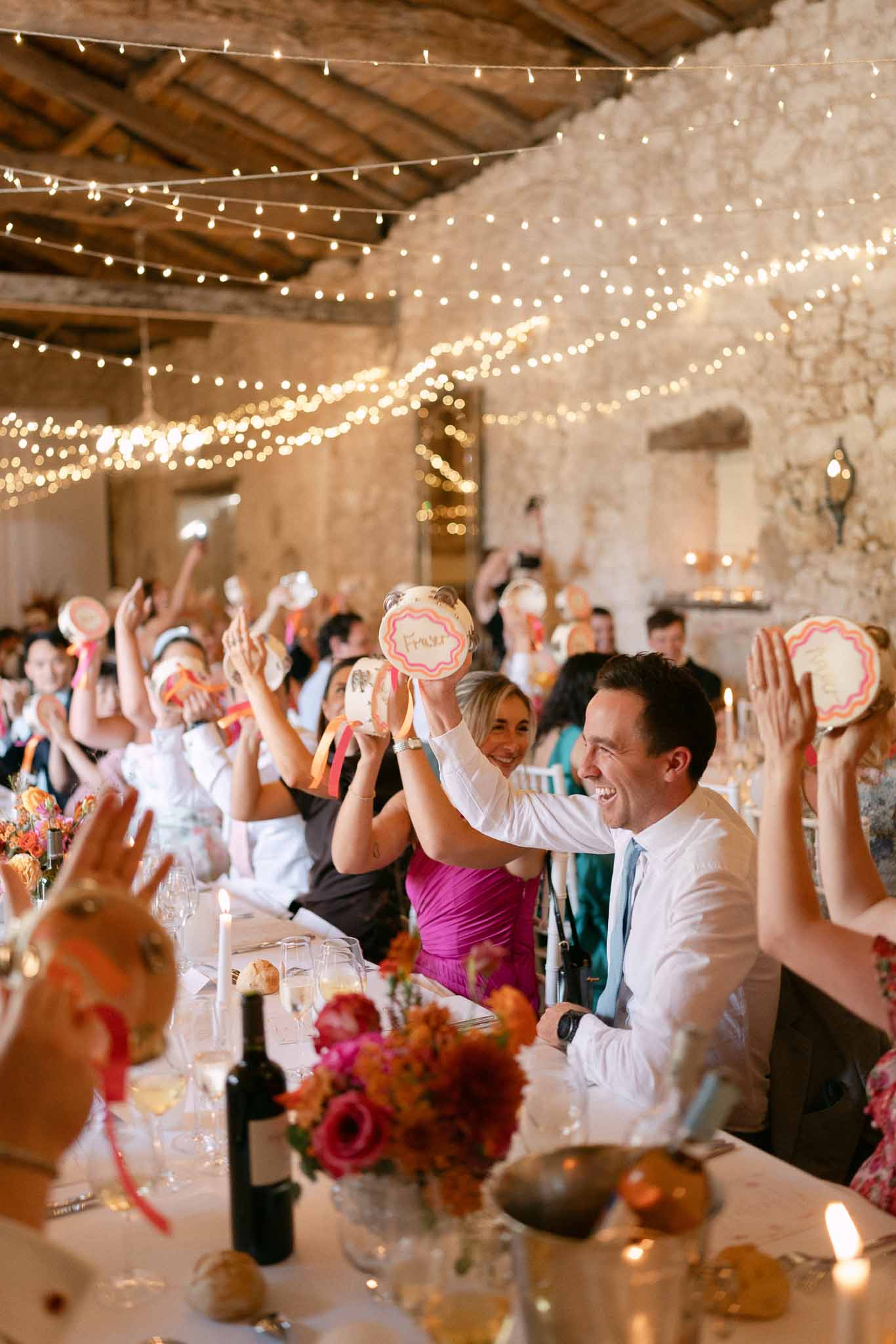 Wedding guests raising decorated tambourines at long tables inside stone barn with fairy lights and colorful floral center...