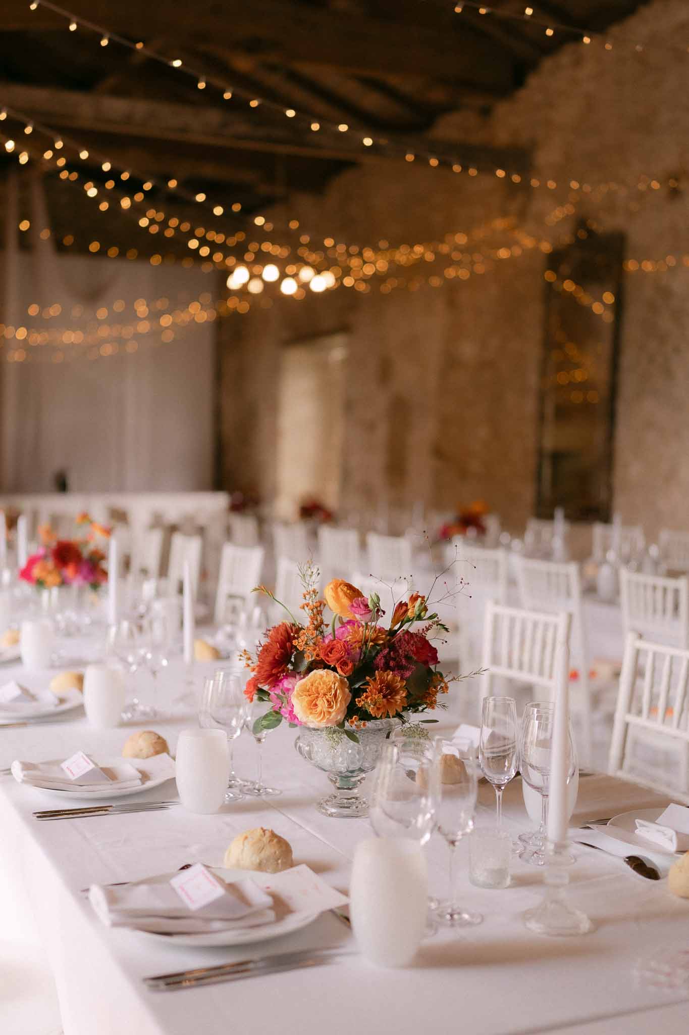 Reception table in stone barn with fairy lights, white linens, and jewel-toned centerpieces of orange roses and burgundy d...