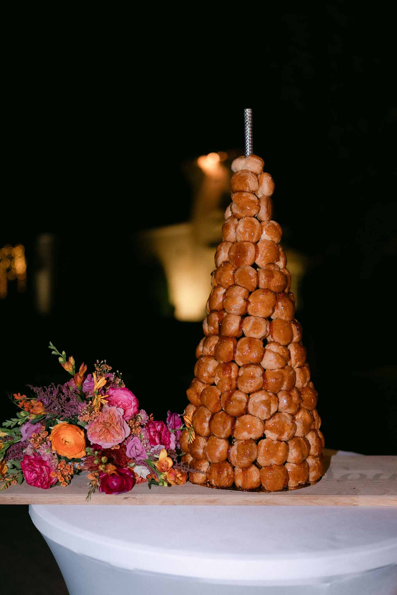 Caramel-glazed croquembouche tower beside hot pink roses and orange ranunculus at evening reception