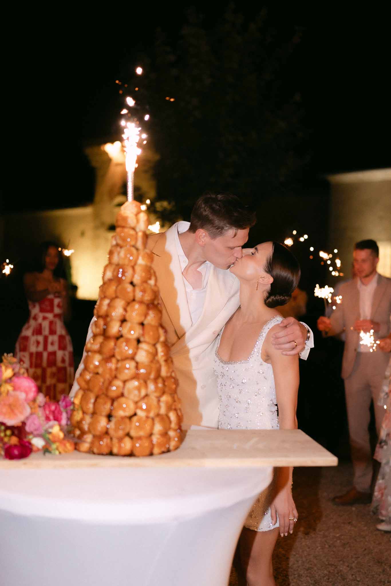 Couple kissing beside croquembouche topped with sparkler at evening reception with hot pink and coral flowers