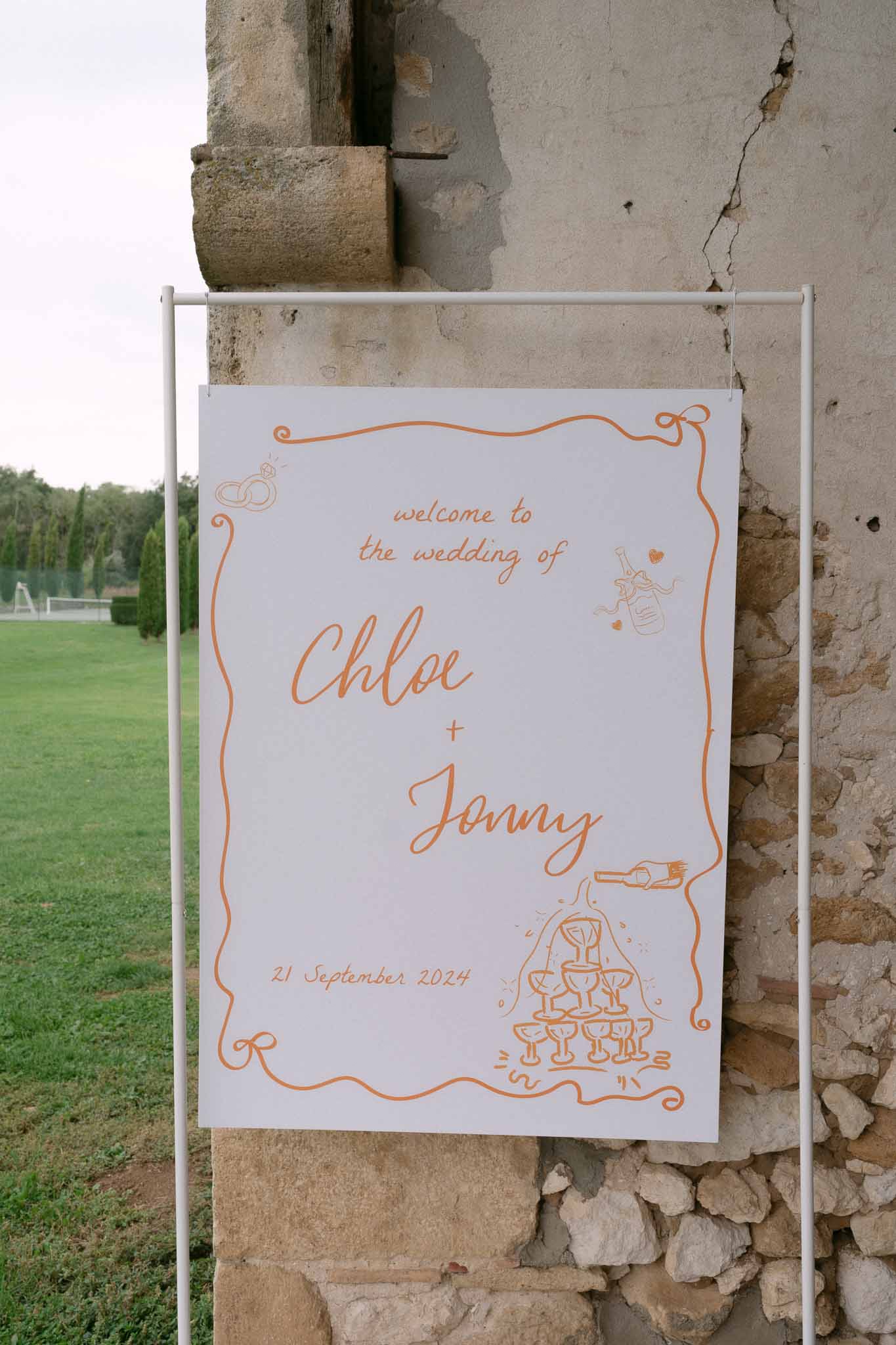 Hand-drawn wedding welcome sign in burnt orange lettering on white board displayed against rustic stone wall