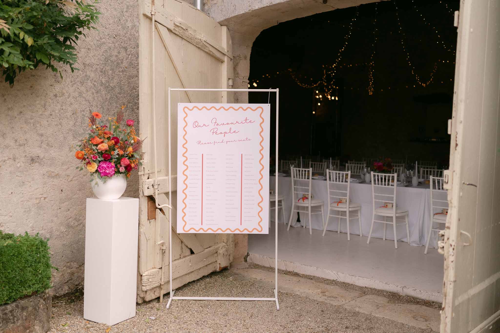 Seating chart sign with pink script and orange border beside hot pink dahlia arrangement at reception entrance