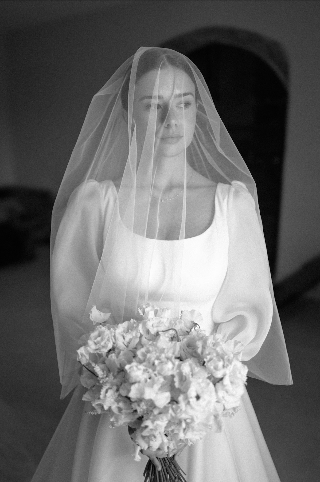 Bride with cathedral veil over face holds dense lisianthus bouquet in chapel with arched backdrop in B&W