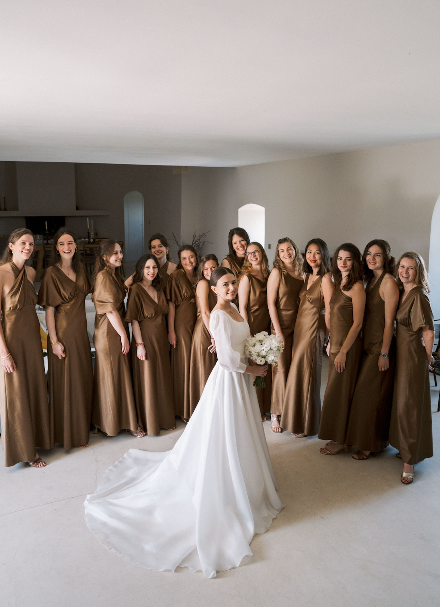 Bride in white gown with 12 bridesmaids in bronze-gold satin dresses in arched stone interior