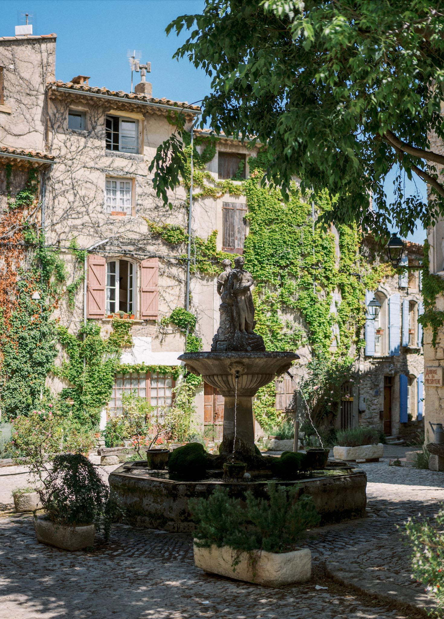 Provencal village square with two-tiered stone fountain, cobblestones, and ivy-covered limestone facades