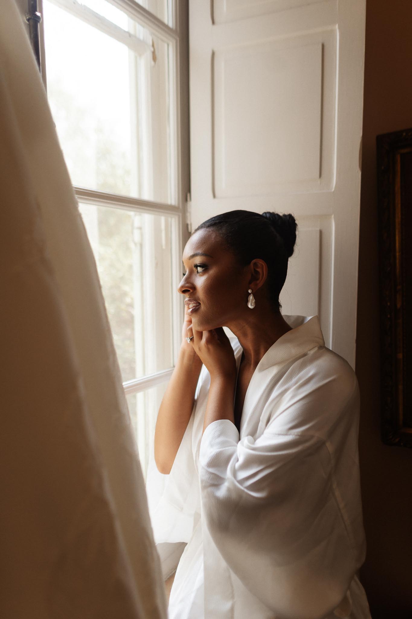 Bride in silk robe looking out window during getting ready preparations in bright interior room