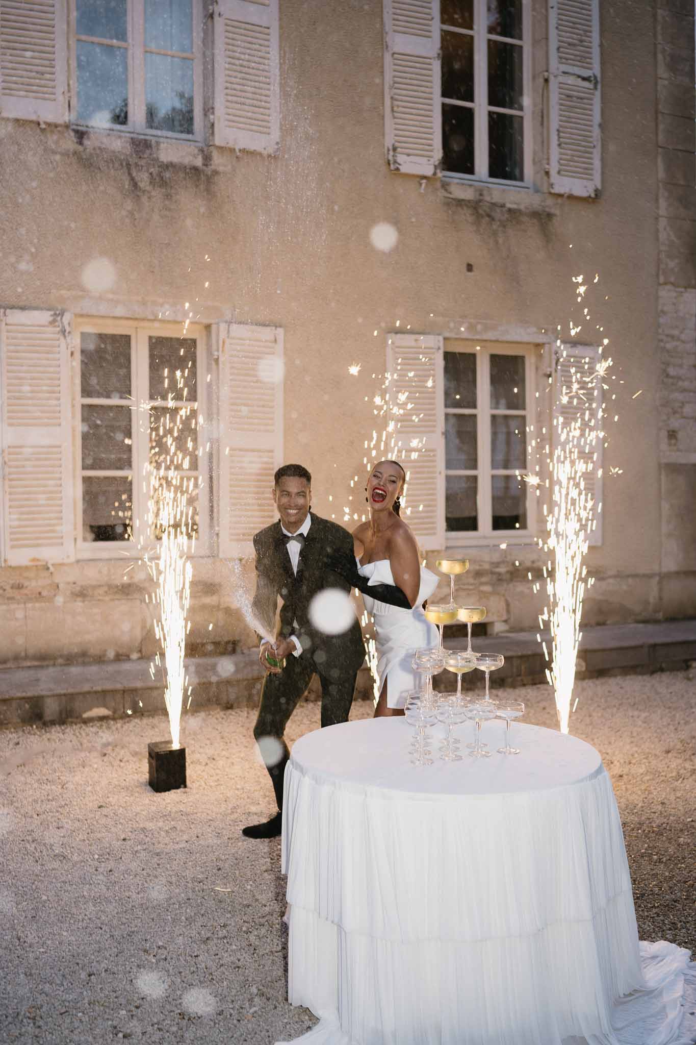 Couple celebrating with confetti during cocktail hour in historic stone courtyard with fairy lights and champagne tower