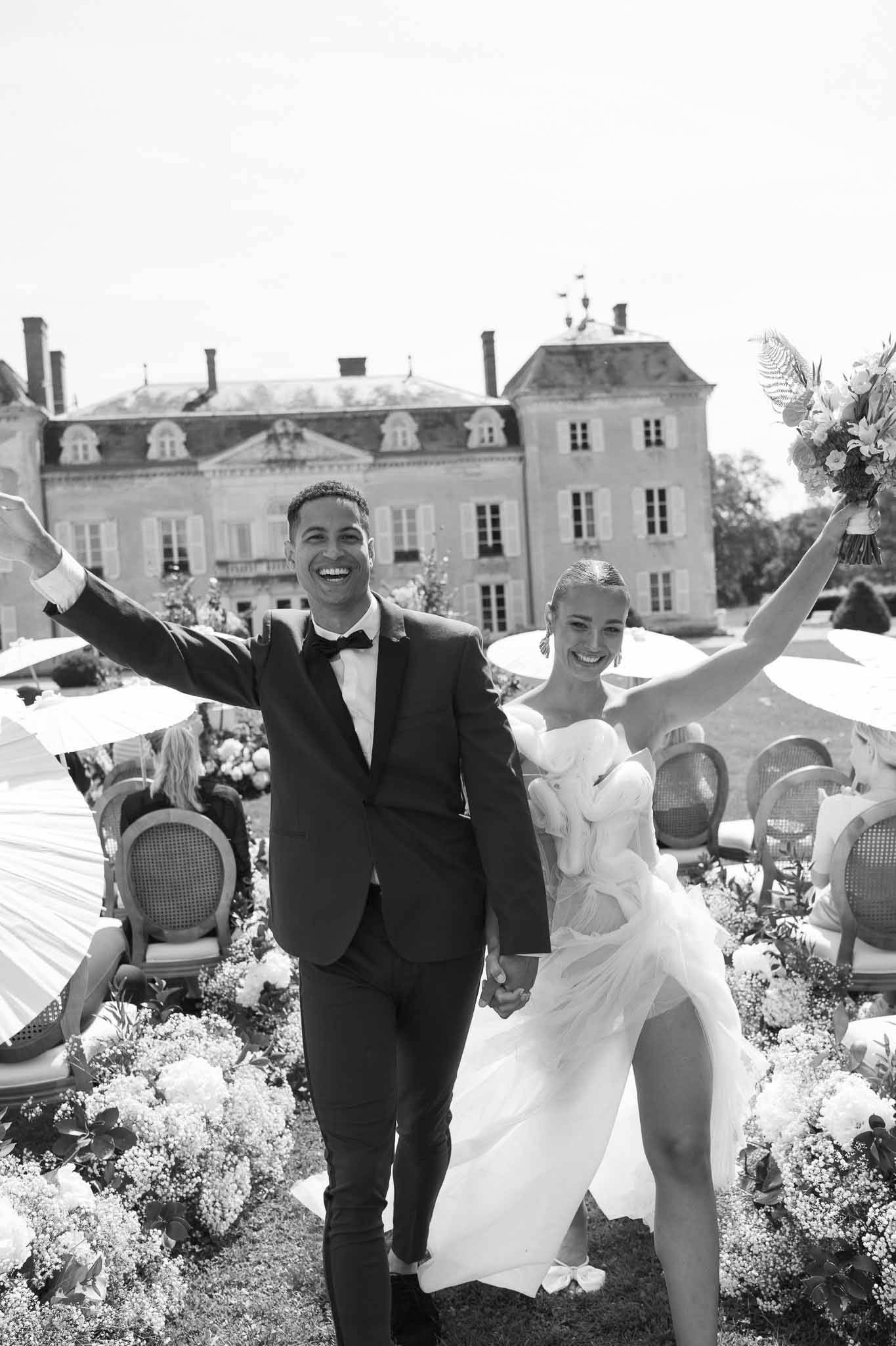 Newlywed couple celebrating at outdoor château terrace reception with guests and floral arrangements